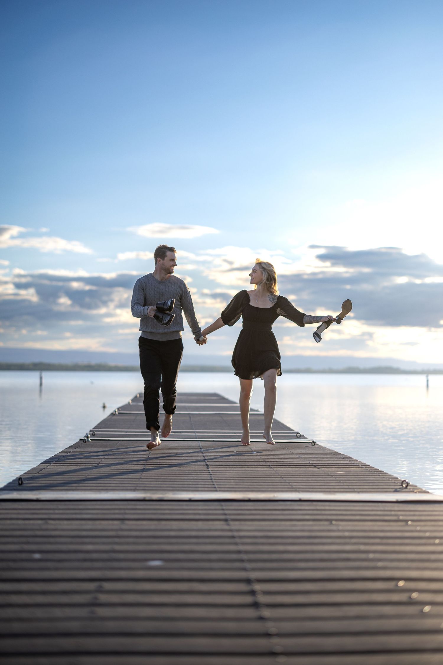 A couple holding hands and running along a wooden pier with shoes in hand during sunset.