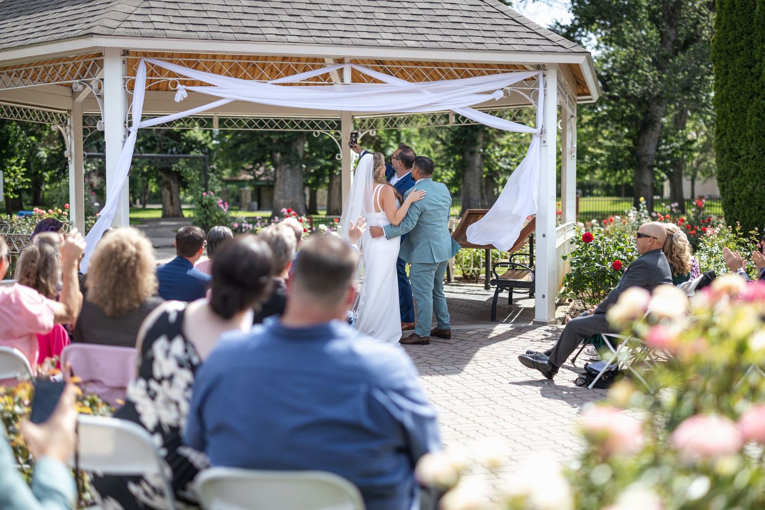 Garden wedding setup features white chairs and gazebo with blooming flowers in foreground.