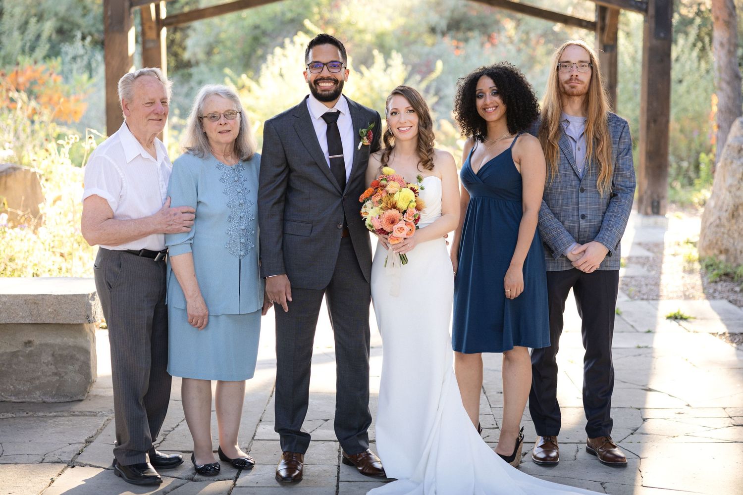 Family members gather for formal wedding portrait under rustic pavilion in garden setting.