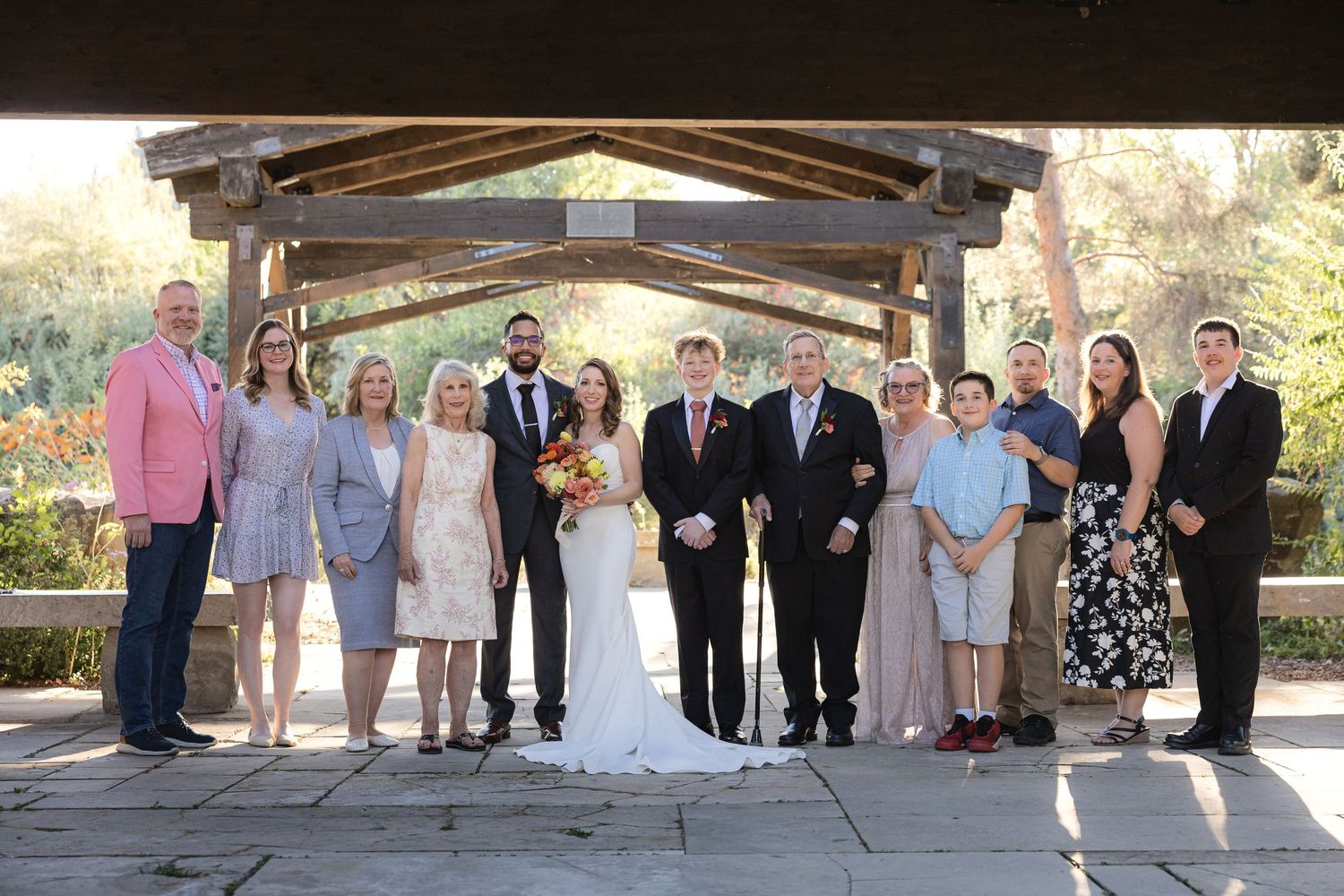 Wedding party gathered under wooden beamed outdoor pavilion.