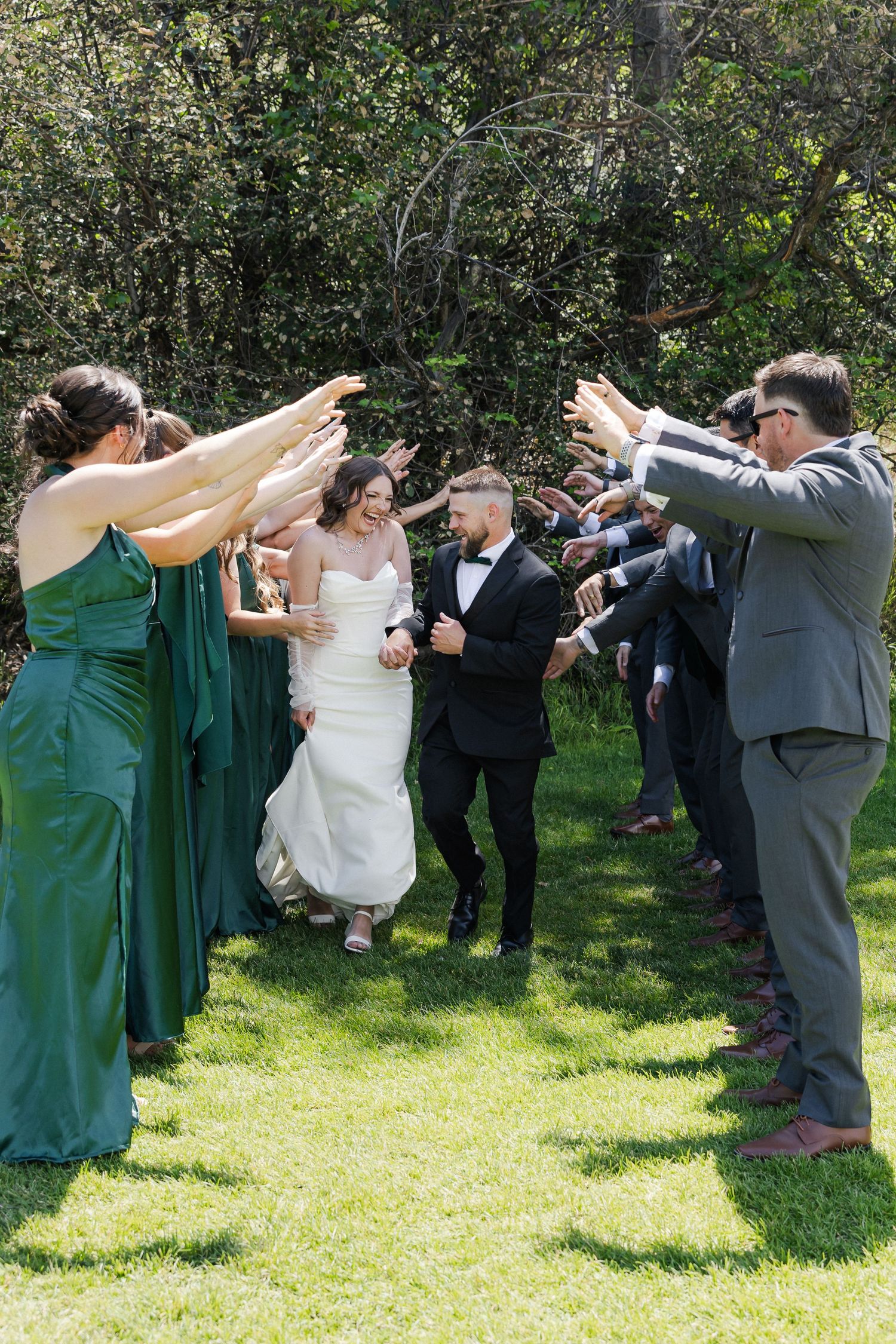Bride and groom walk through a celebratory arch of hands formed by wedding party in green and gray attire on a sunny lawn.
