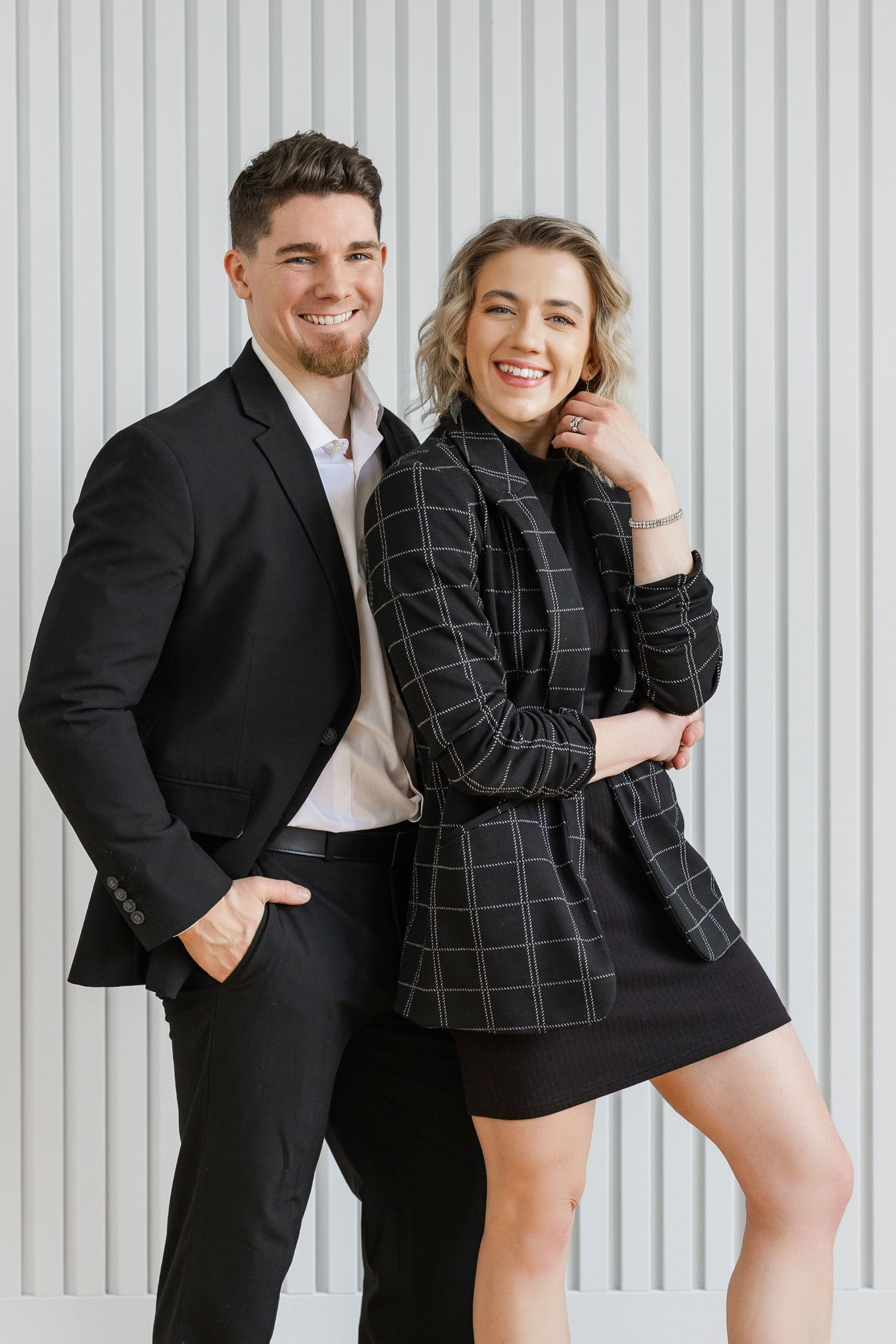 Playful couple in formal attire posing against white striped wall background.