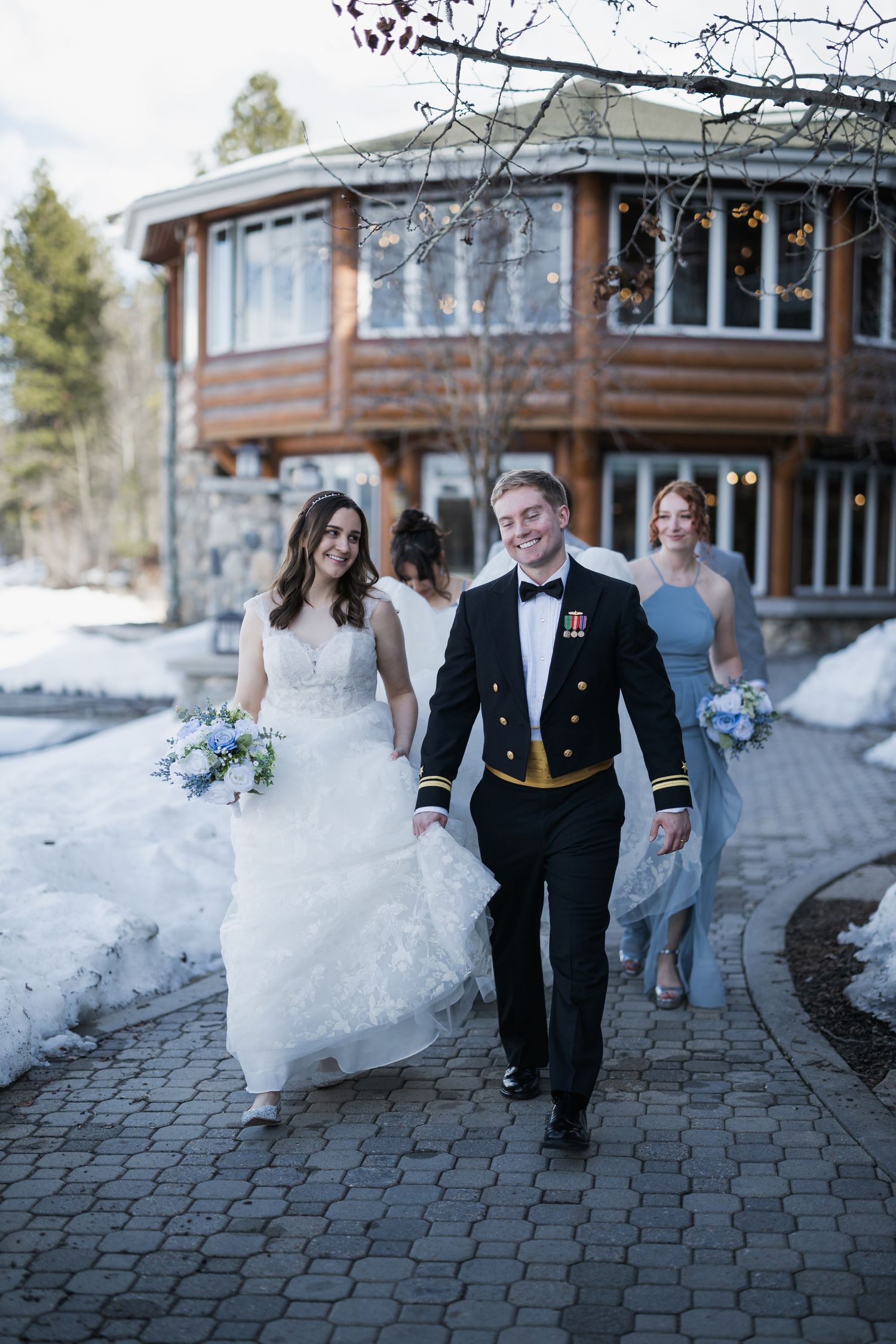 A wedding couple and wedding party walk along a snowy cobblestone path outside a rustic lodge venue in winter.