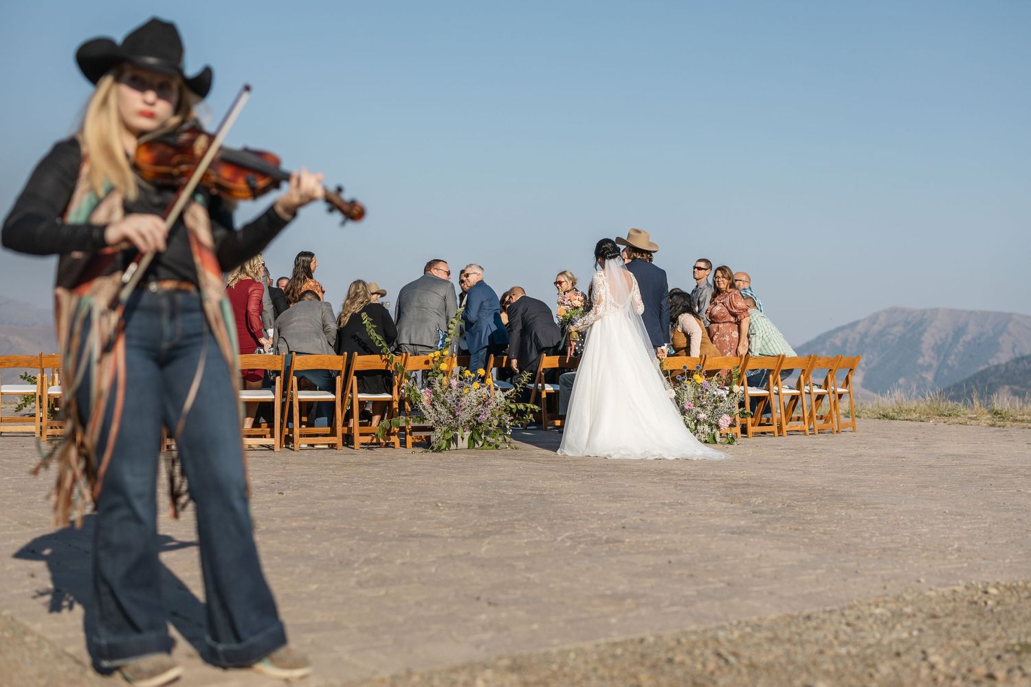 Outdoor mountain wedding ceremony with violinist playing and guests seated on wooden chairs.
