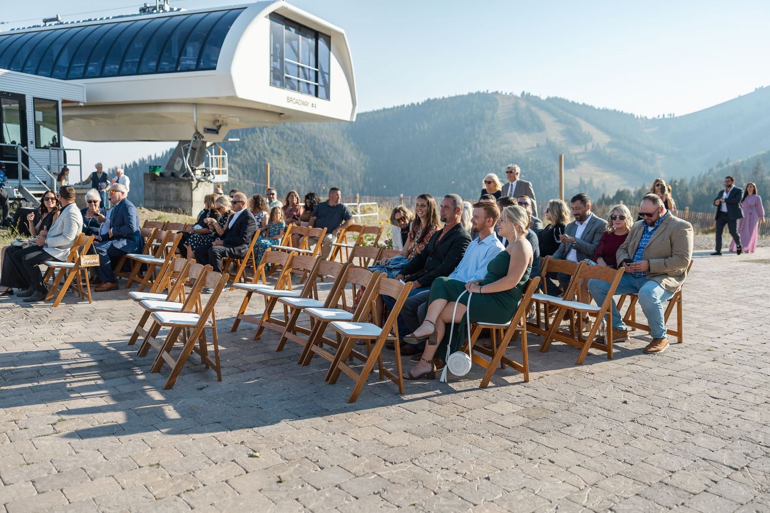 Wedding guests seated in wooden folding chairs on a mountain summit with scenic views and a modern venue structure.