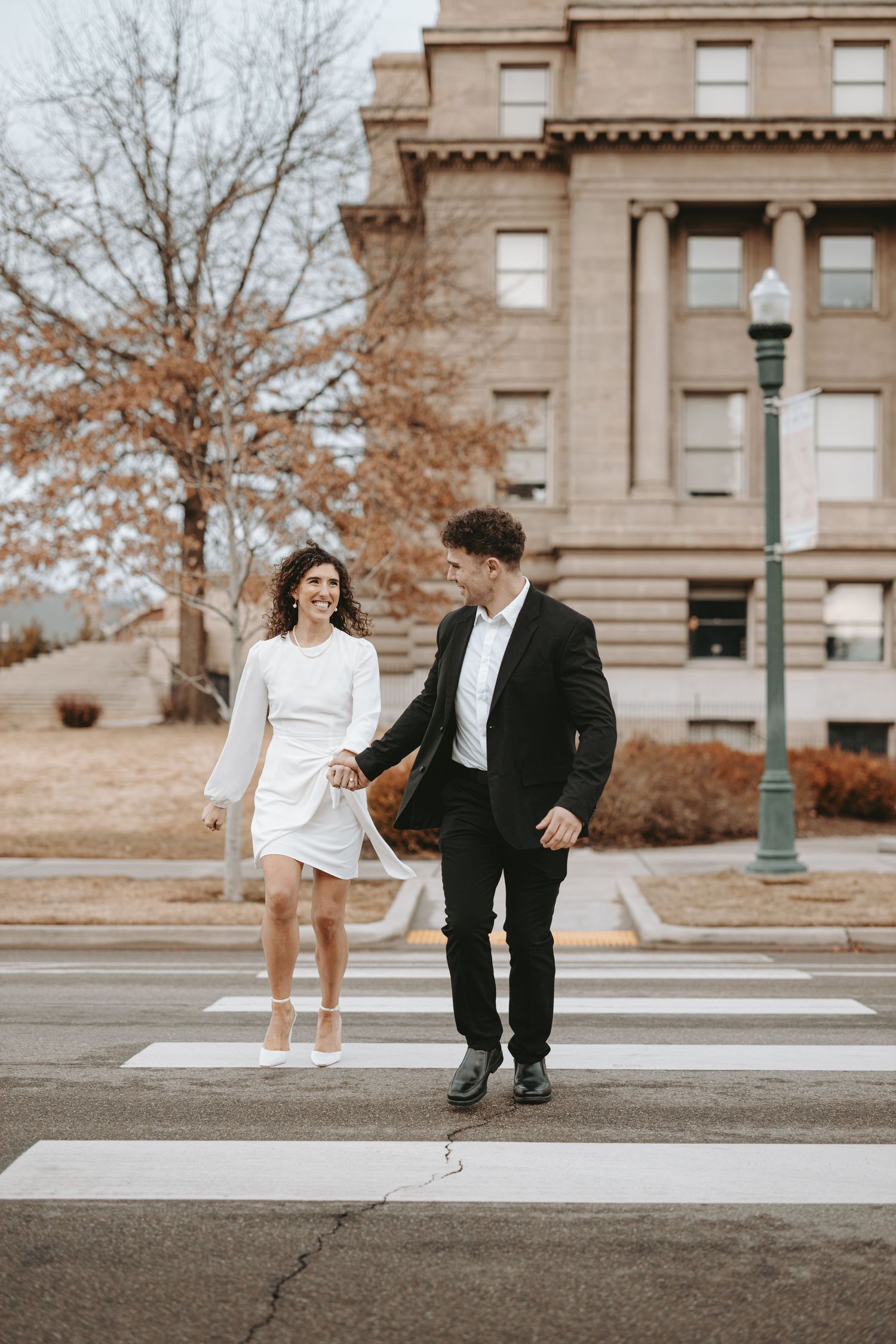 A couple holds hands while crossing a street in front of a stately building with autumn trees.