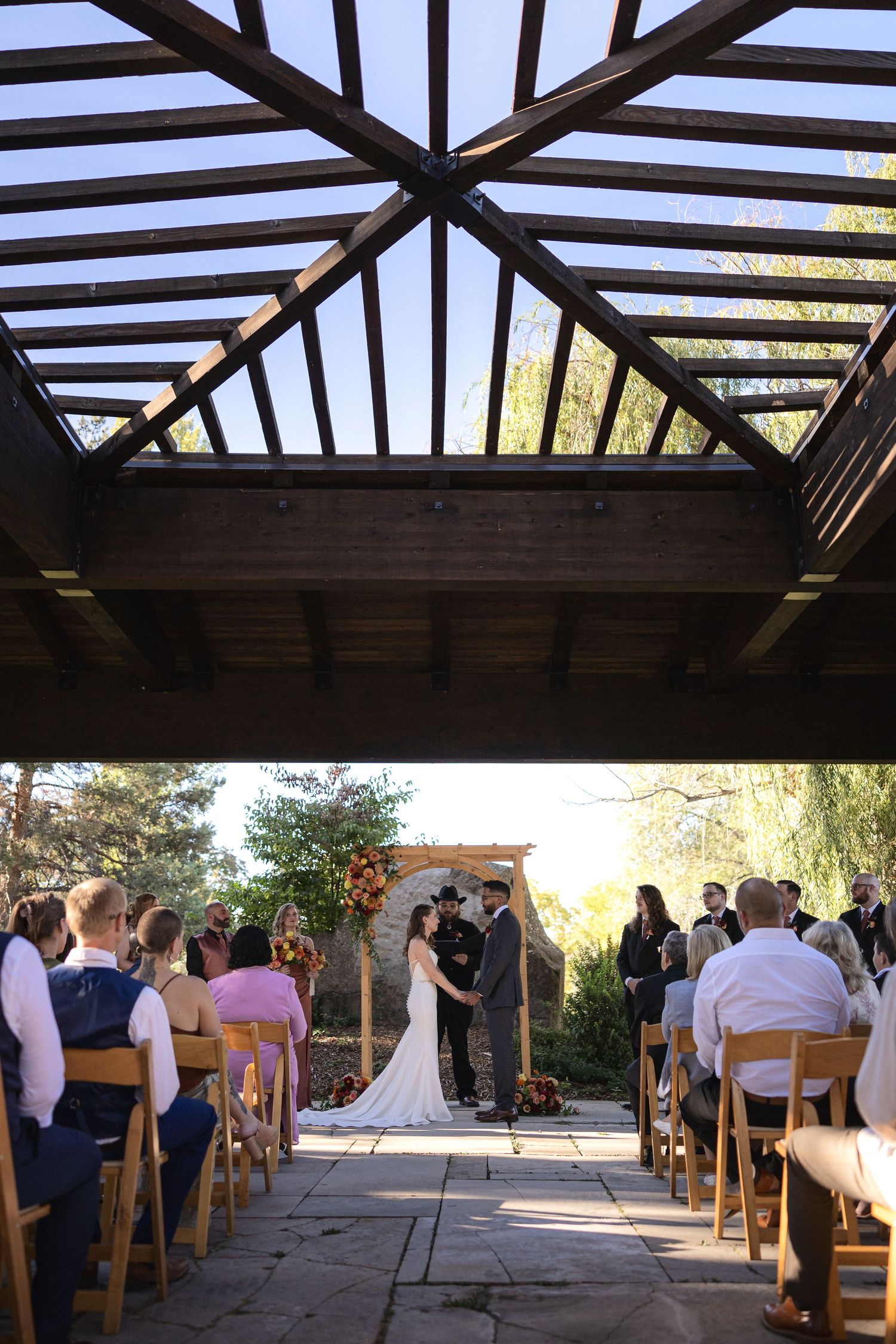 Wedding ceremony viewed through wooden beams of covered pavilion with folding chairs.