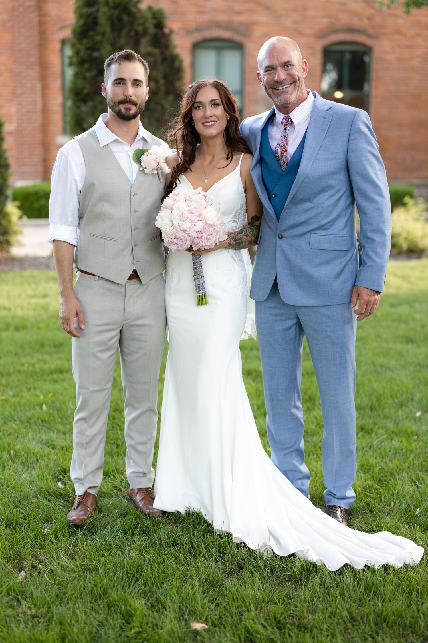 Three people in formal attire pose for wedding photo on lawn with brick building backdrop.