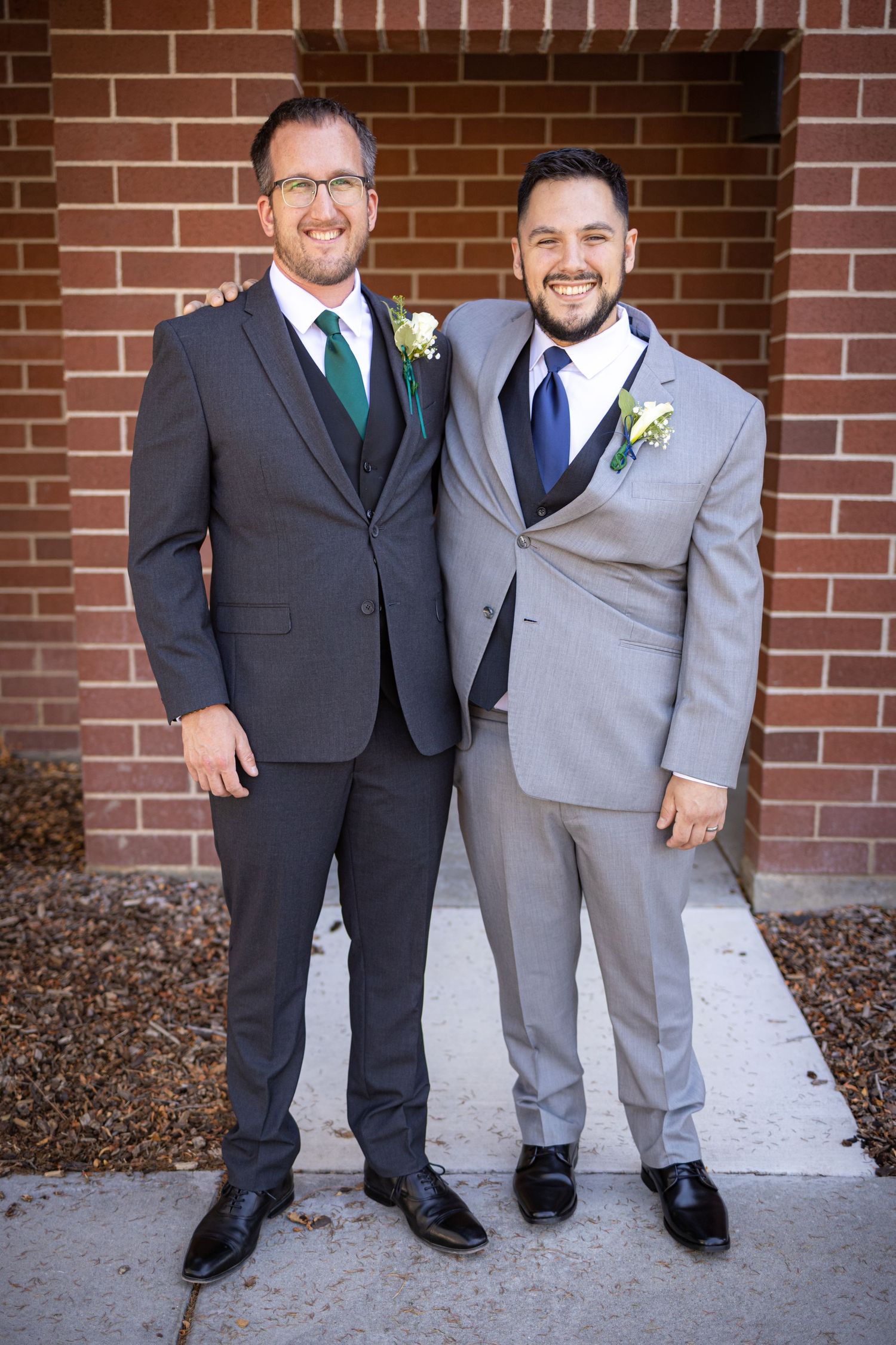 Two men in formal suits stand together against brick wall wearing floral boutonnieres.