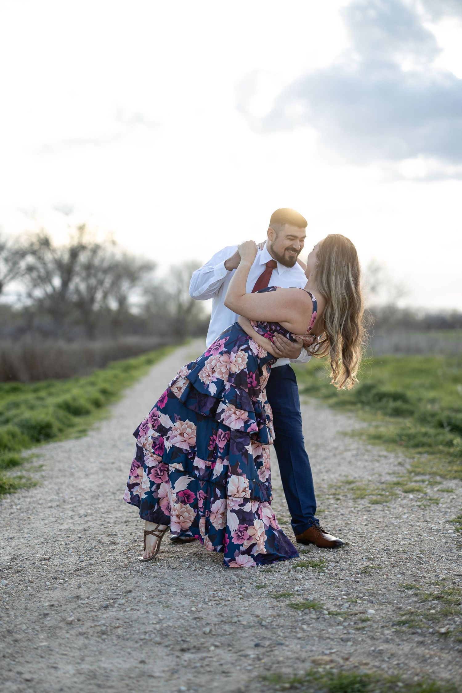 A romantic couple dances on a dirt path surrounded by green fields at sunset.