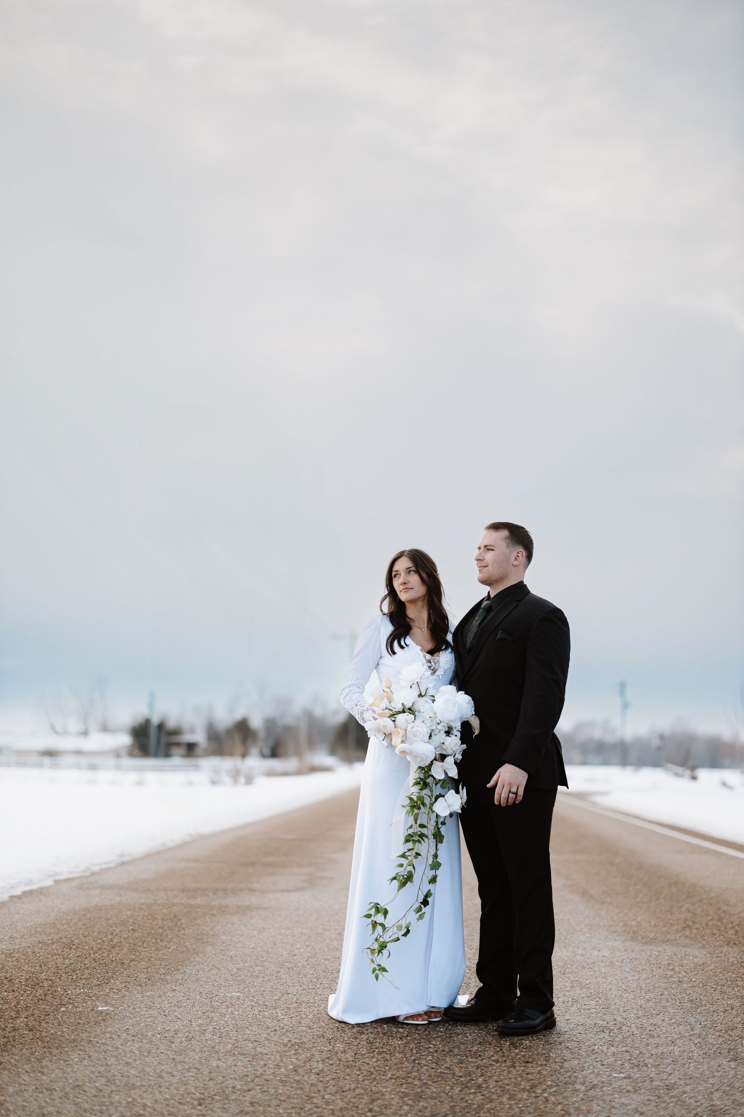 A formal couple poses on a snow-lined road with a cascading white floral bouquet.