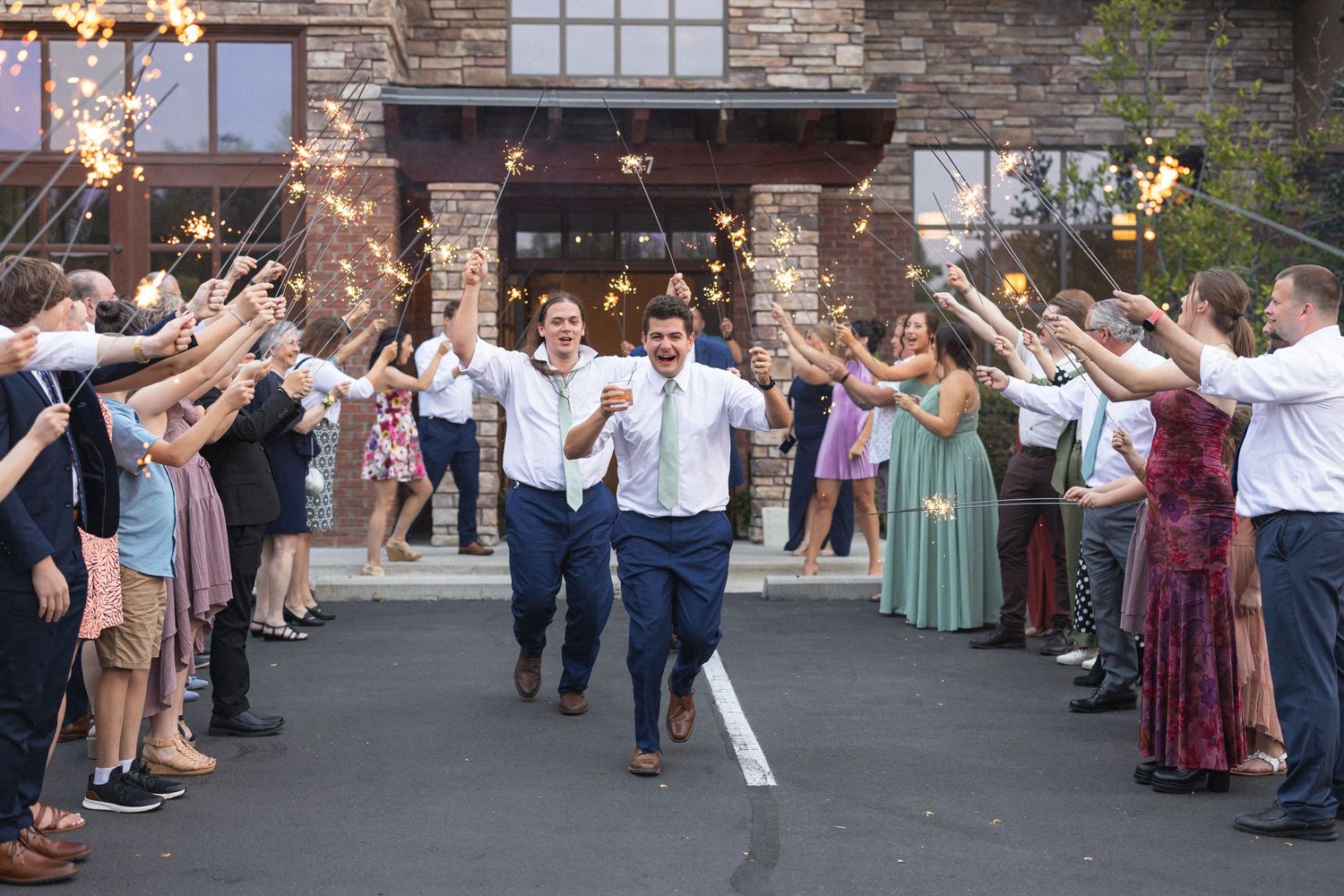 Wedding guests create a sparkler send-off tunnel for couple exiting stone building at night.