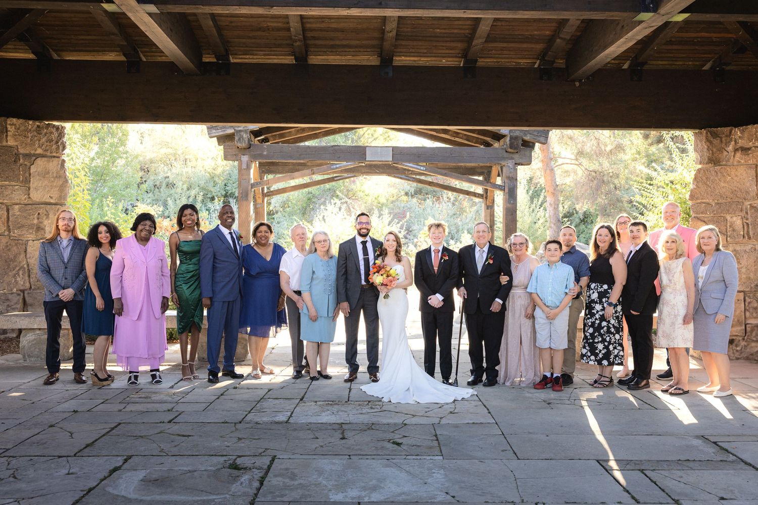 Large wedding party group photo under rustic covered pavilion with stone architecture.