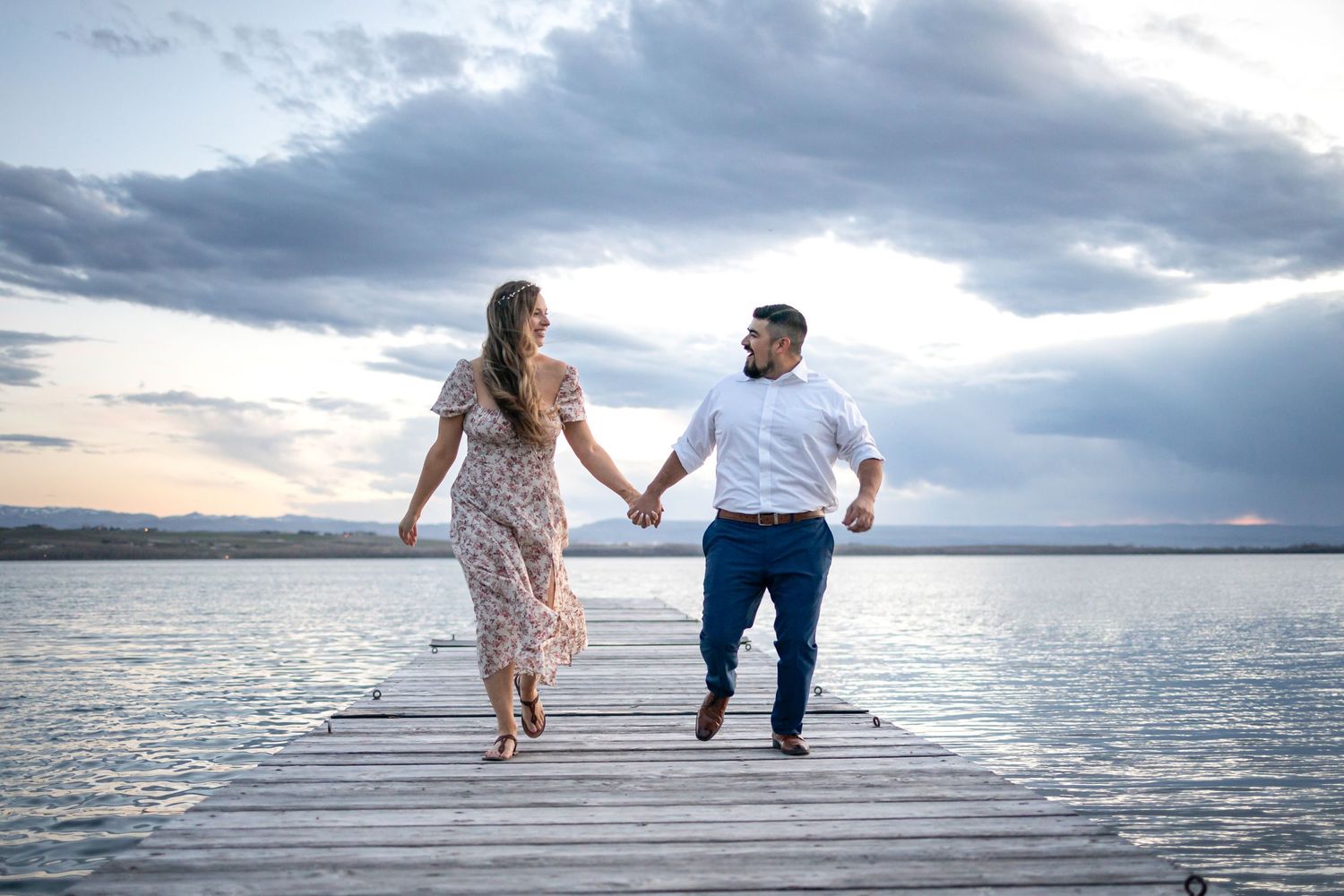 A romantic couple walks hand in hand on a wooden dock over a lake during sunset with dramatic clouds.