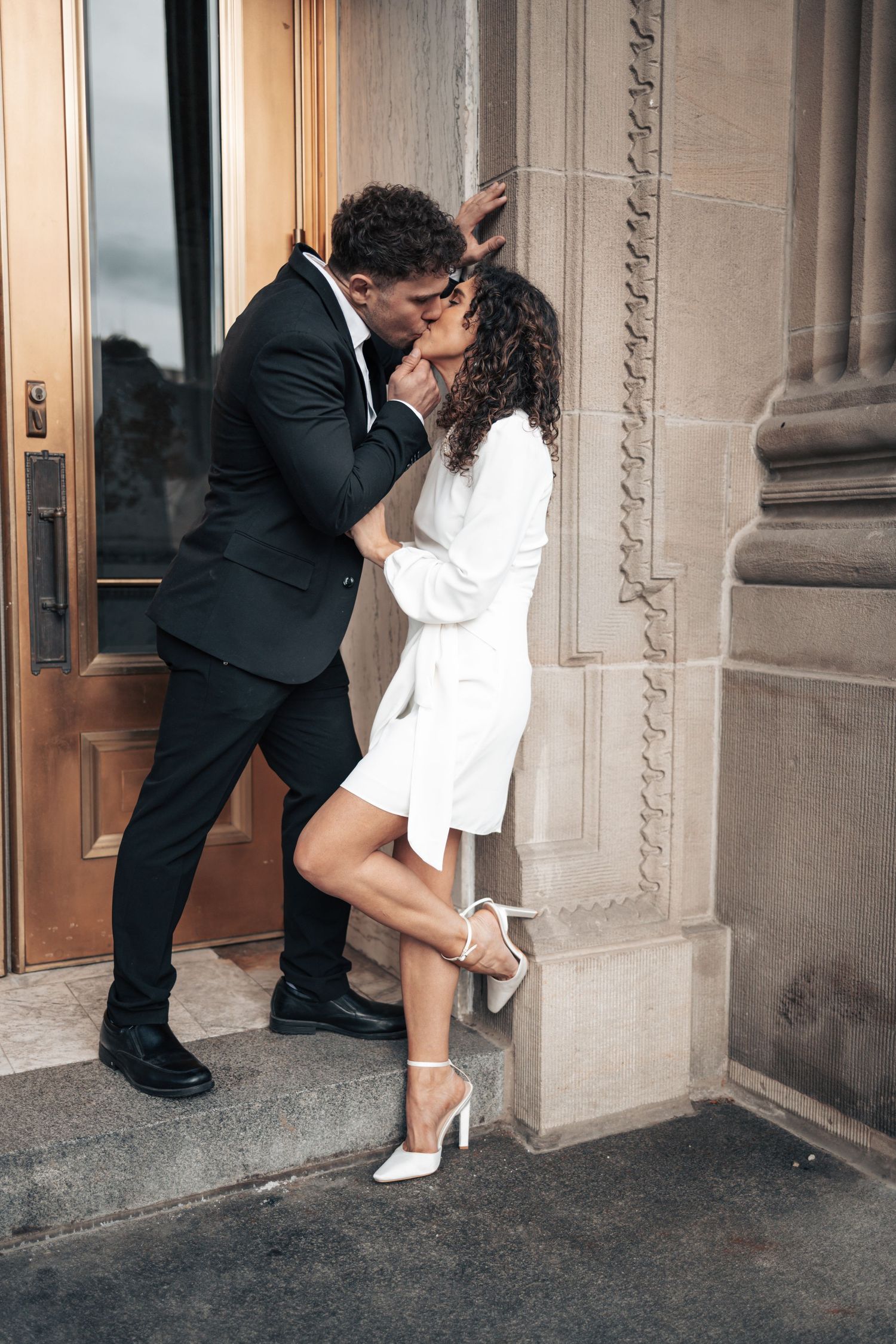 A romantic couple in formal attire share a passionate kiss against a stone building with elegant doors.