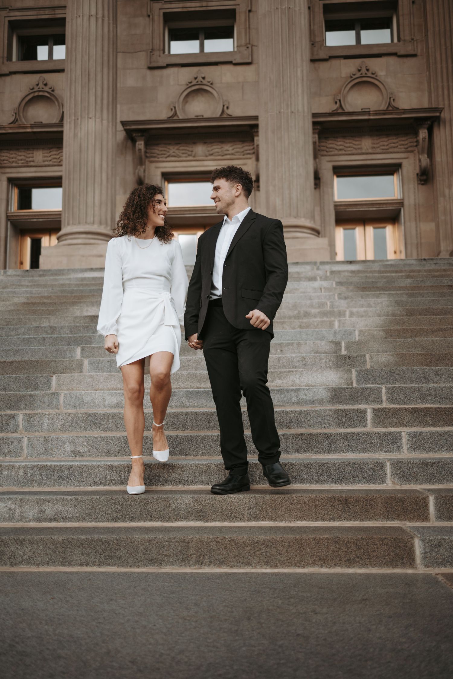 A couple in formal wear stands together on stone steps of a grand building.