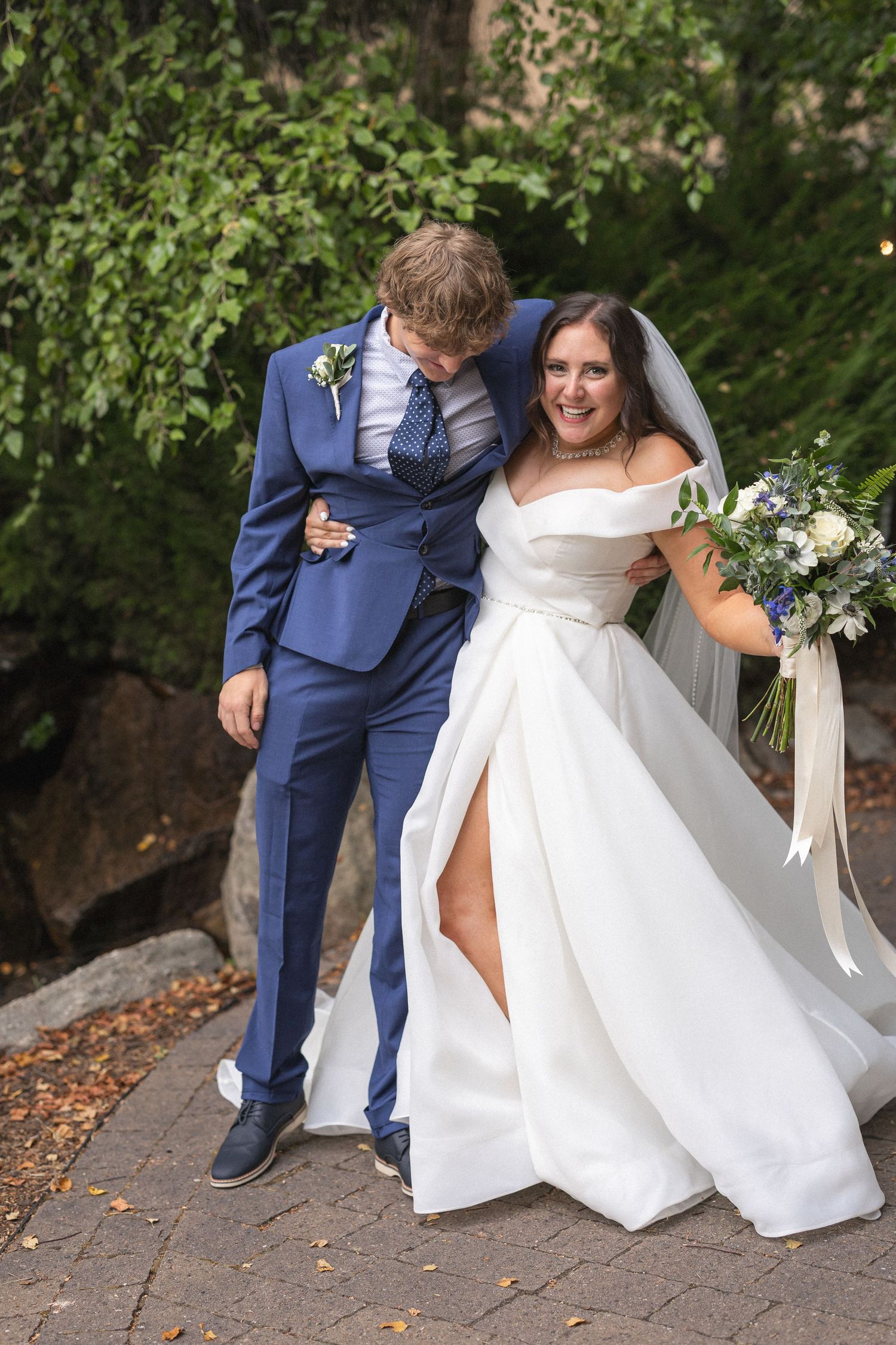 Wedding couple laughs together on stone path, bride in off-shoulder gown and groom in blue suit.
