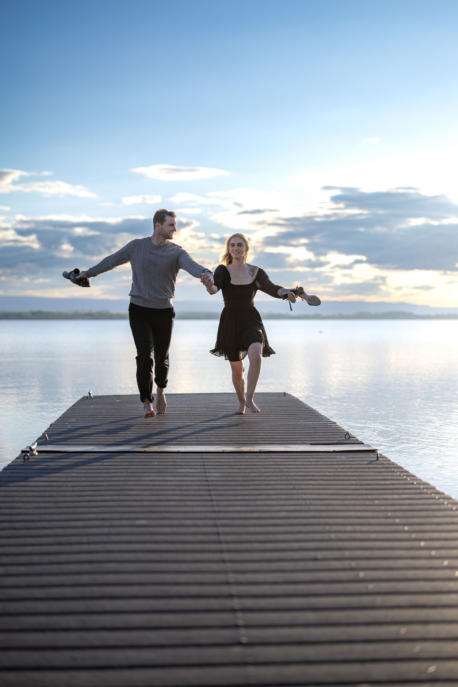 Two people dancing joyfully on a wooden dock with their shoes in hand during sunset.
