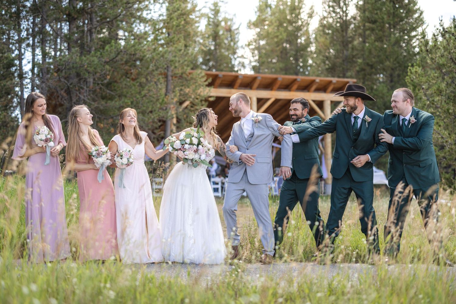 Wedding party poses together in front of rustic wooden structure with evergreen trees in background.