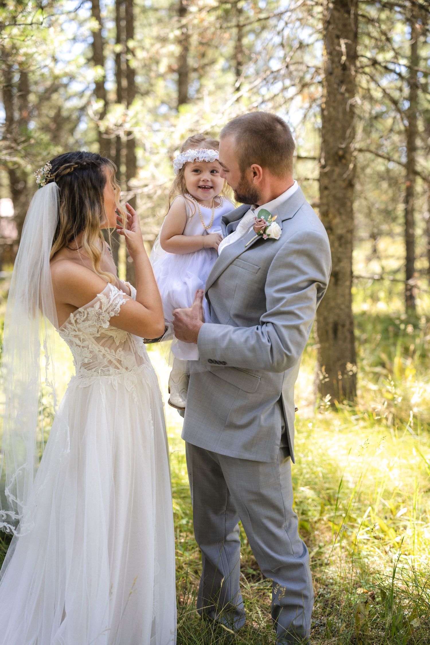 Wedding couple shares joyful moment with young flower girl in sun-dappled forest setting.