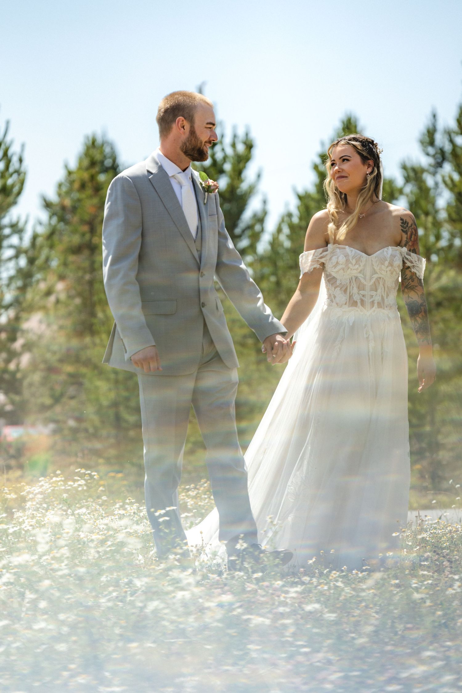 A romantic wedding couple stands together in a field of wildflowers with evergreen trees in the background.