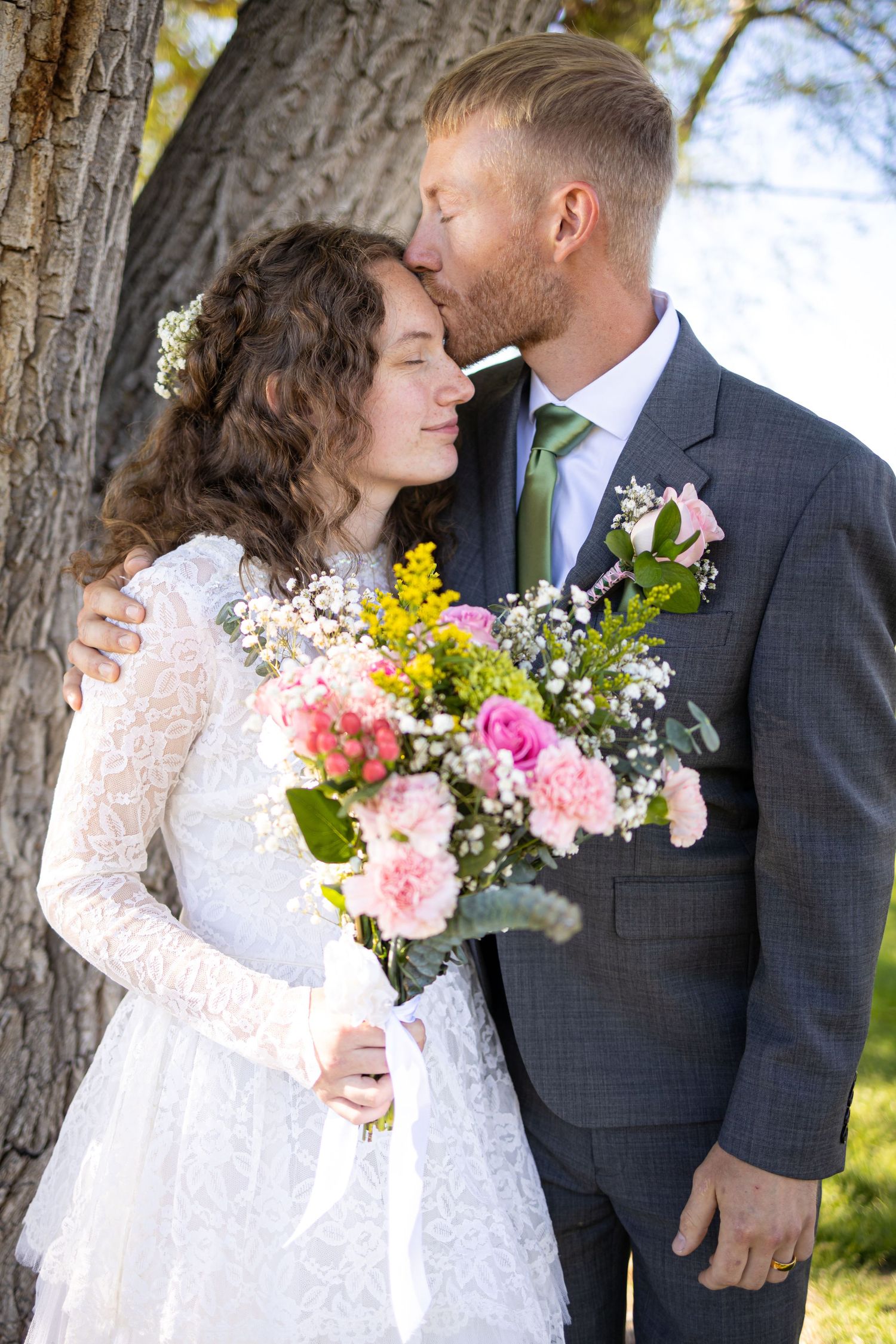 A tender moment between newlyweds standing by a tree trunk with a colorful spring bouquet.