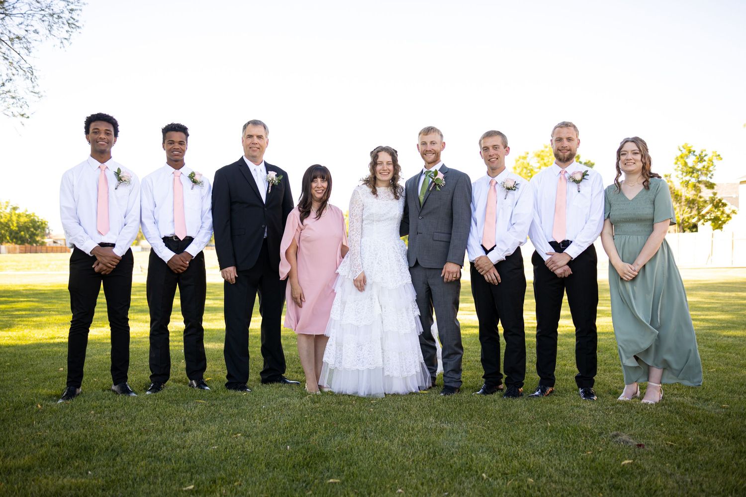 Wedding party poses together on sunny lawn in formal attire with bridesmaids in pink and sage green dresses.
