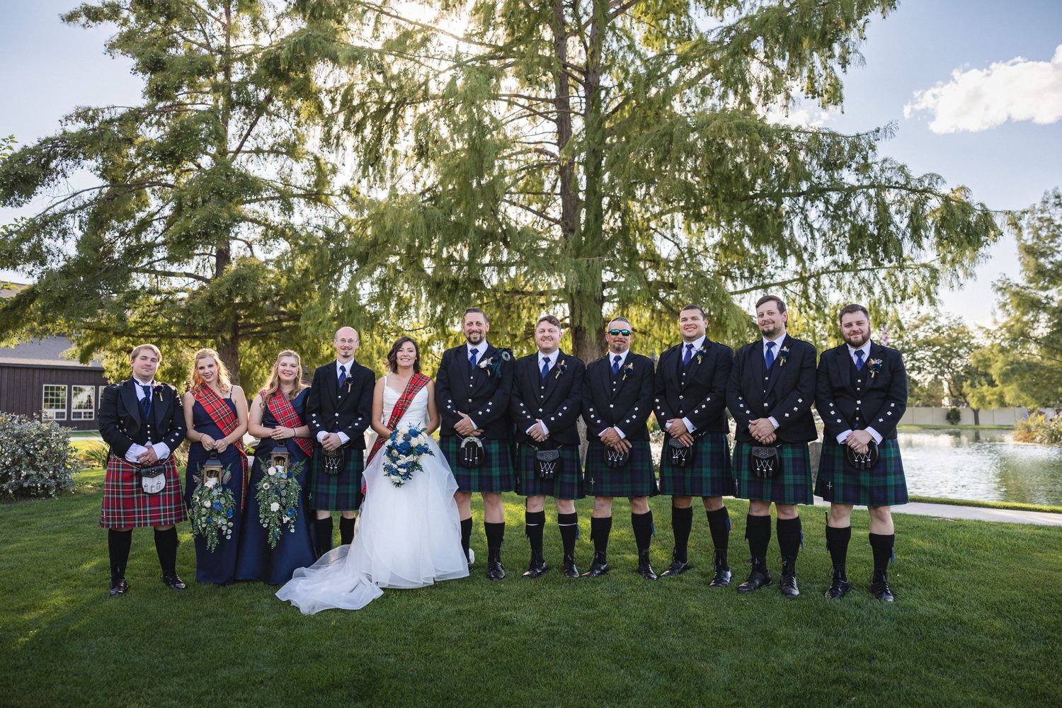 Wedding party lines up on lawn with groomsmen in kilts and bridesmaids in colorful dresses.