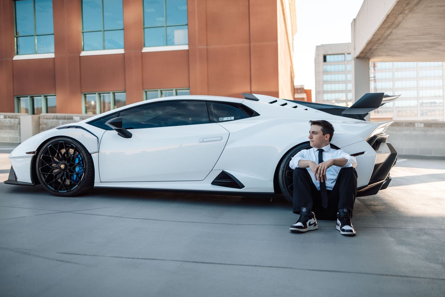 Person in formal wear sitting against white Lamborghini in modern parking garage.