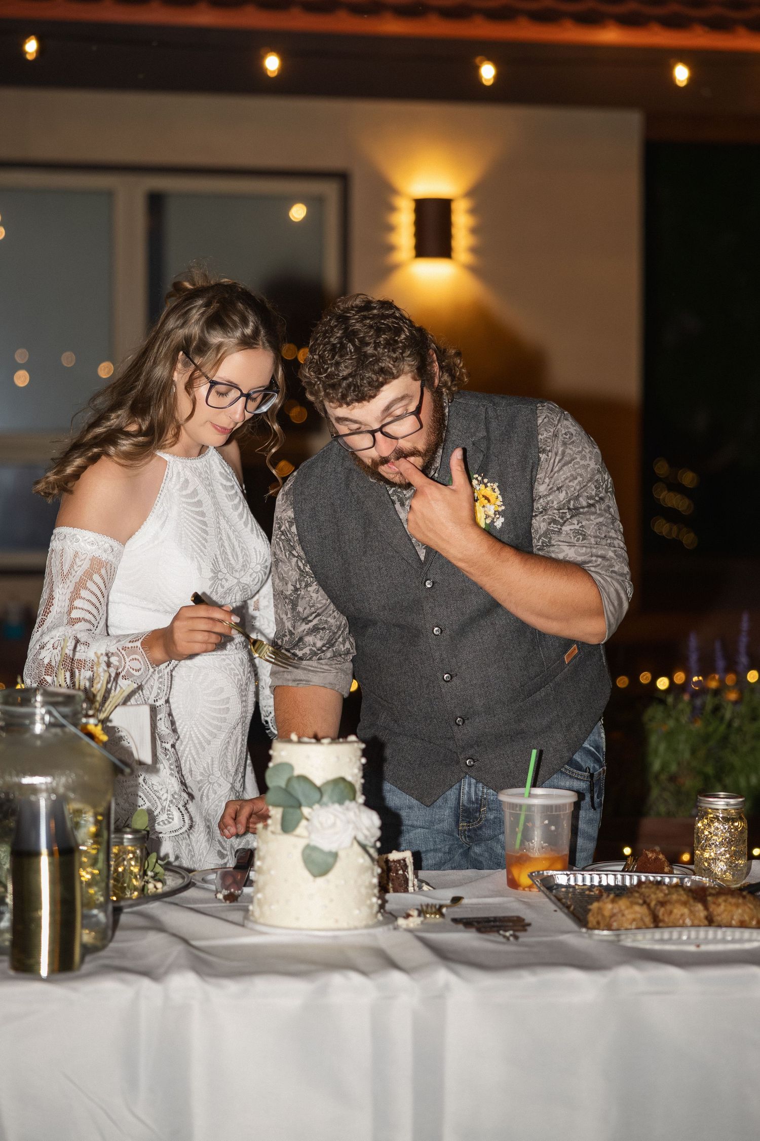 Wedding couple cutting their white wedding cake together at reception with string lights in background.