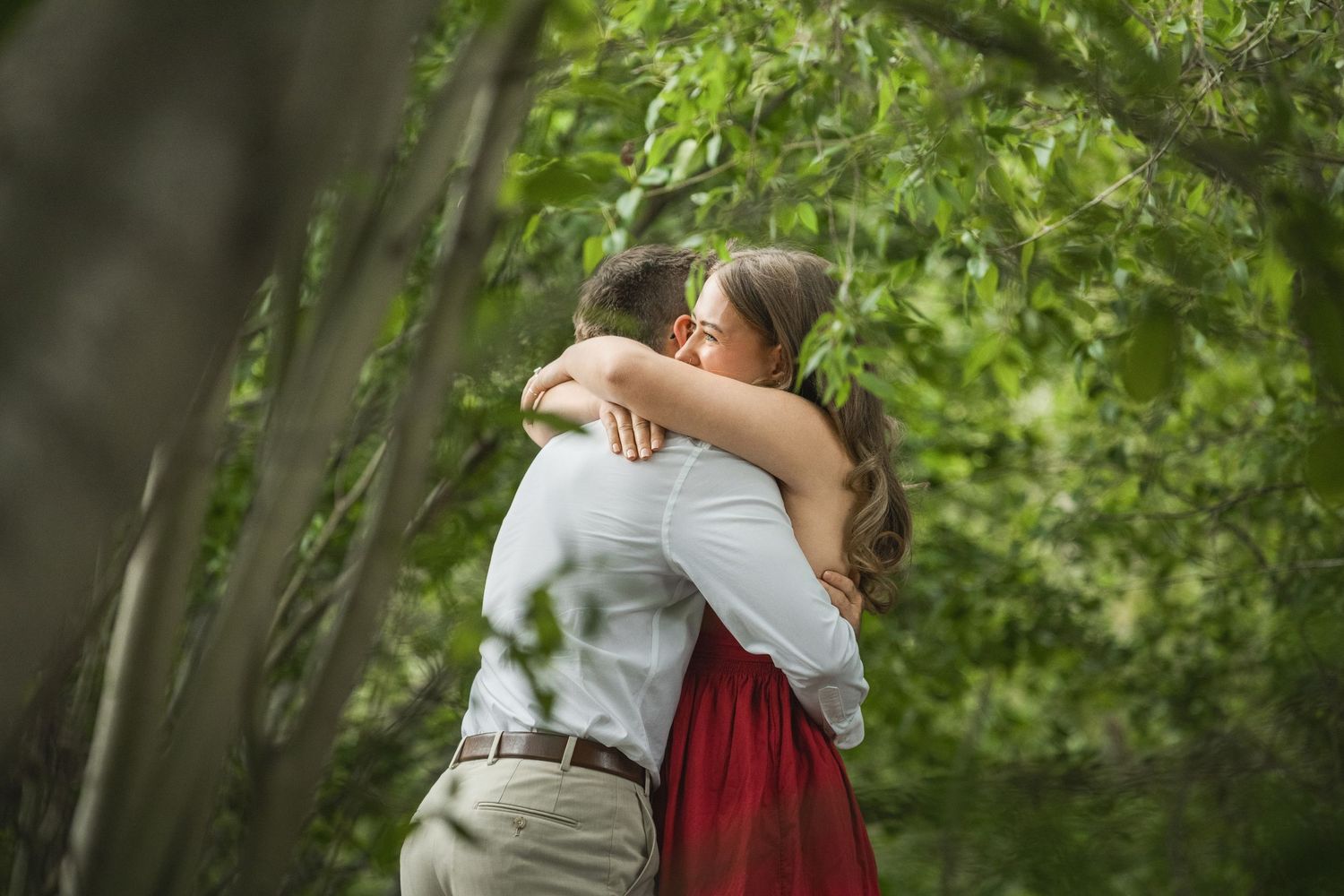 A couple in a forest embrace each other wearing red and grey formal attire during an intimate photo session.