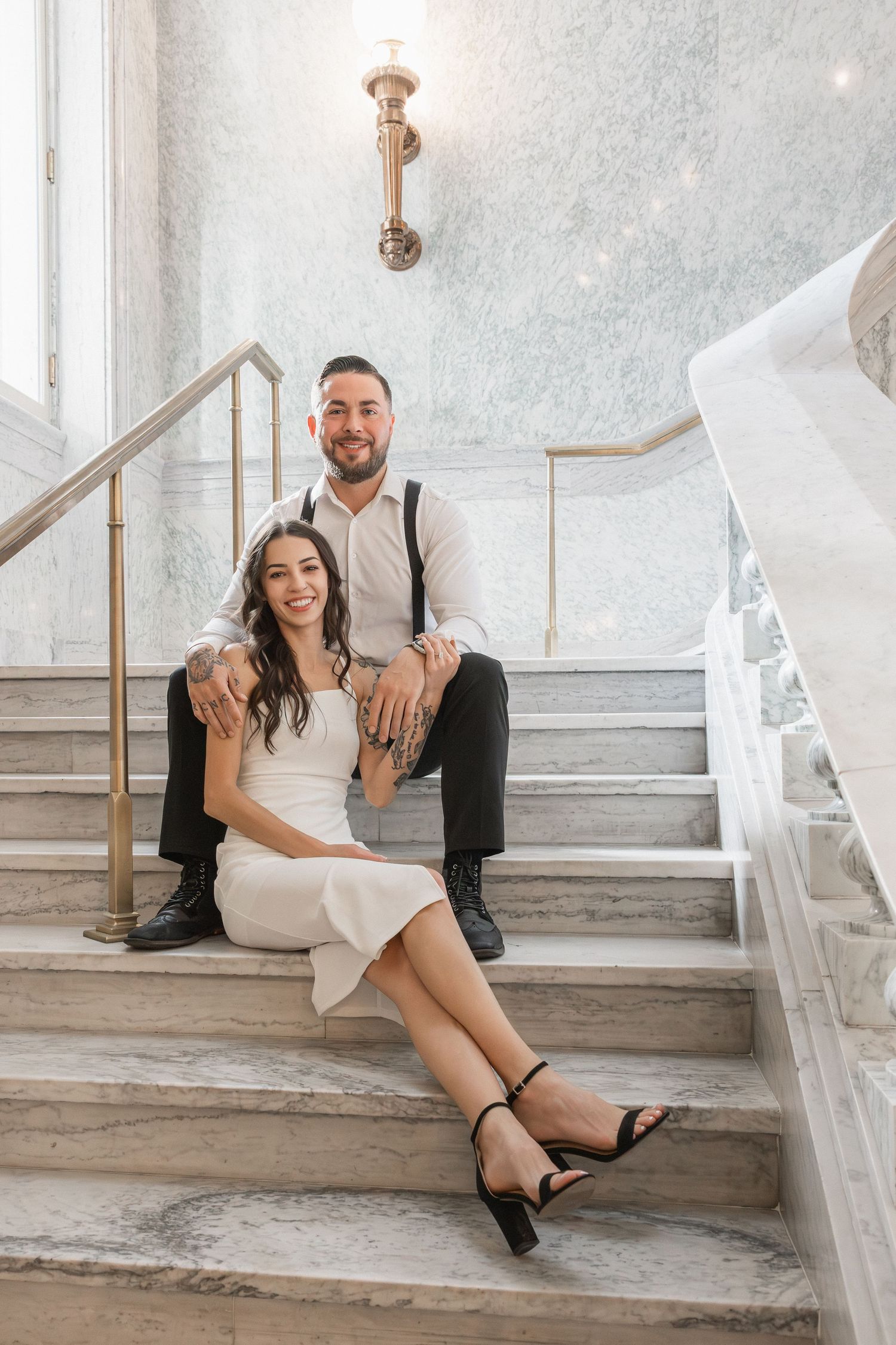 A couple sits casually on elegant marble stairs in a bright interior space with white walls and gold railings.