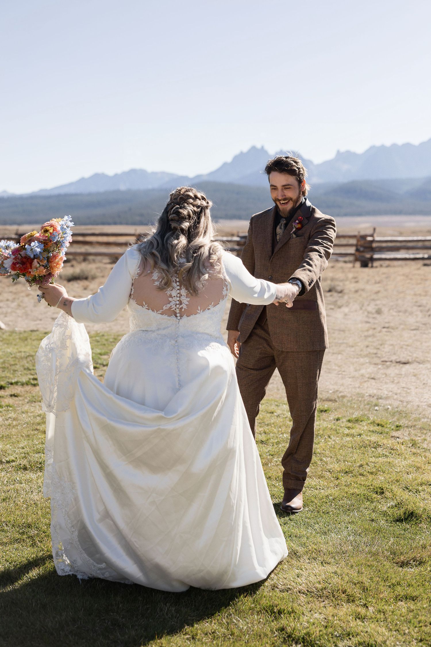 A bride in a flowing white dress twirls in a field with mountains in the background.