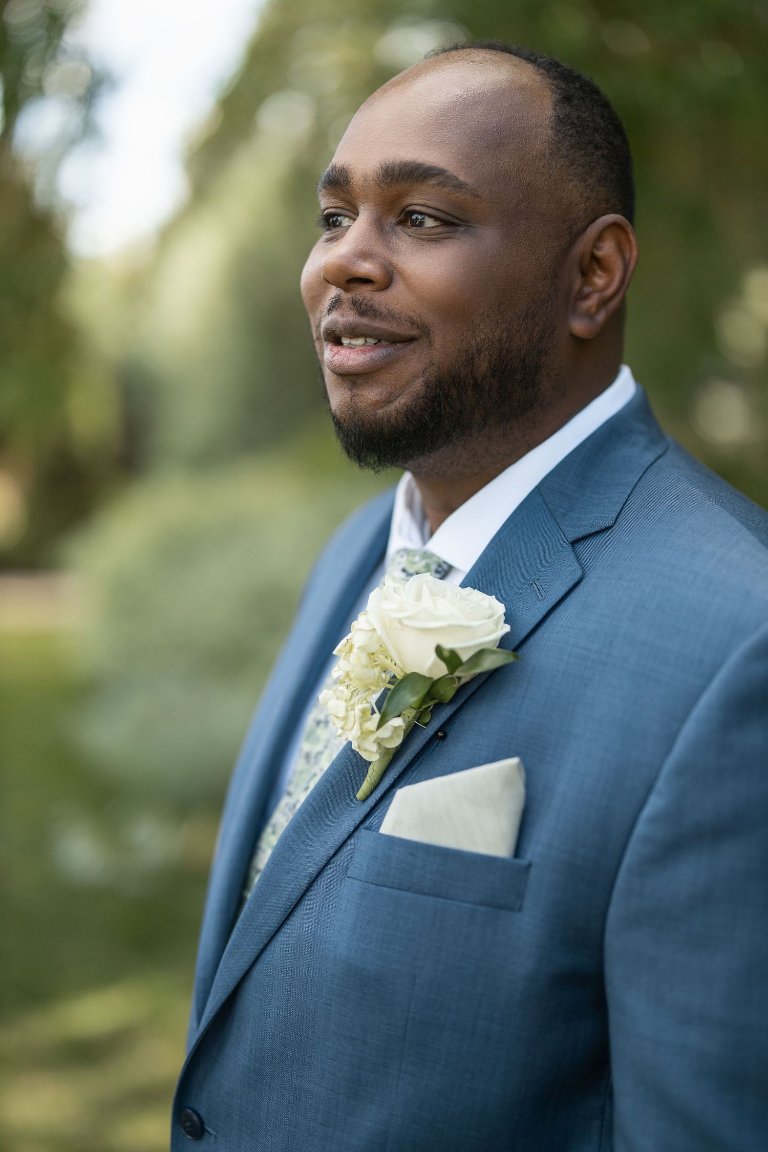 Groom in blue suit with white boutonniere smiling in outdoor setting.
