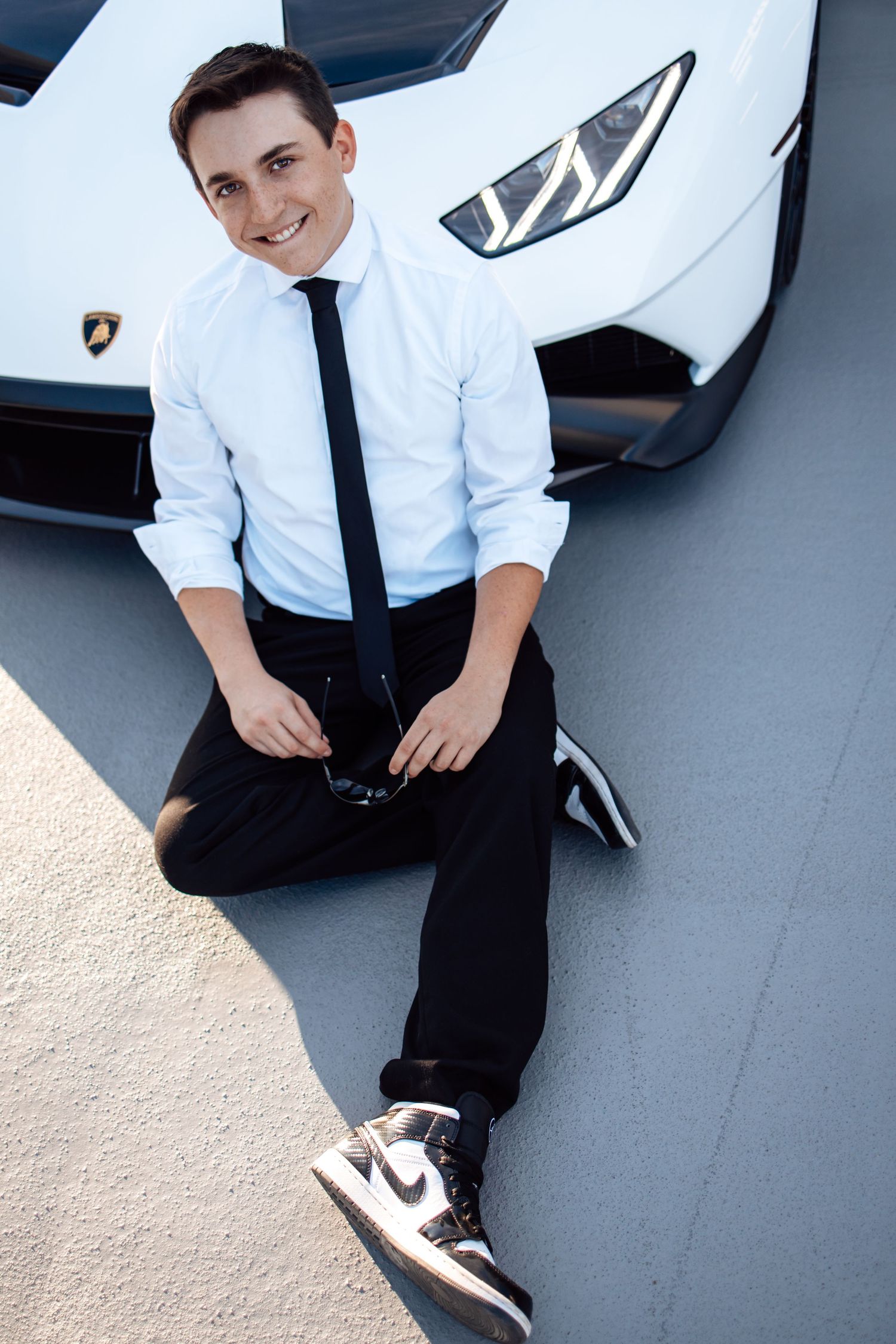 Person in business attire sits casually next to luxury car in parking garage.
