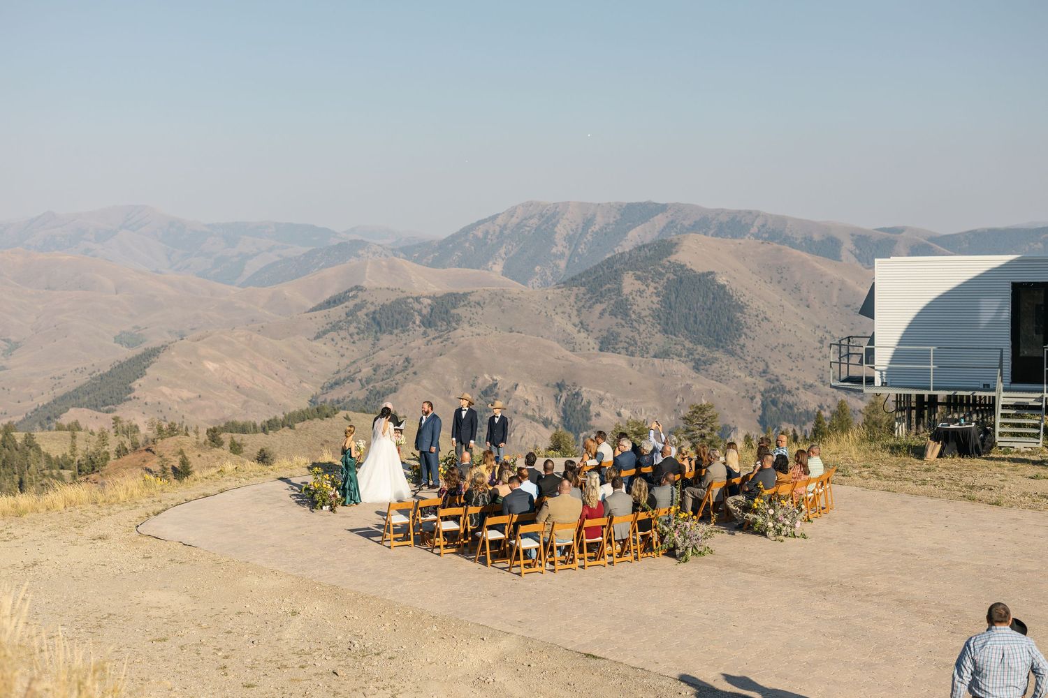 A mountain wedding ceremony site with wooden chairs and guests gathered on a scenic overlook.