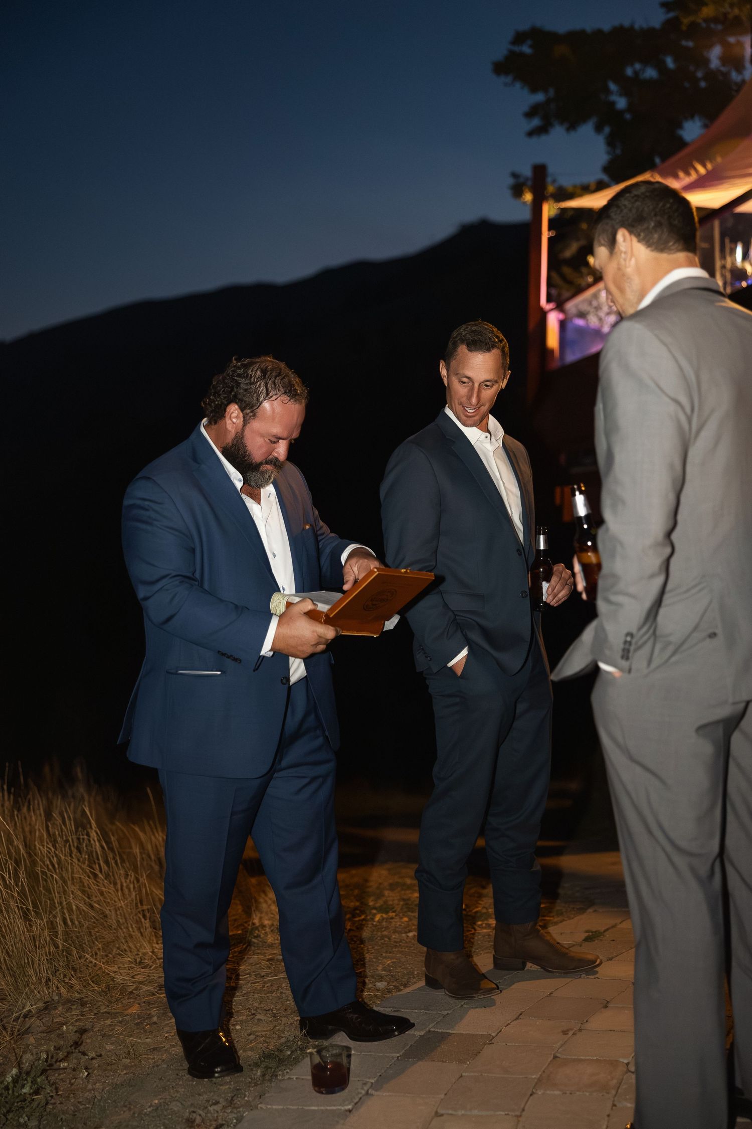 Groomsmen in navy suits and grey suits lighting cigars at an outdoor evening wedding reception.