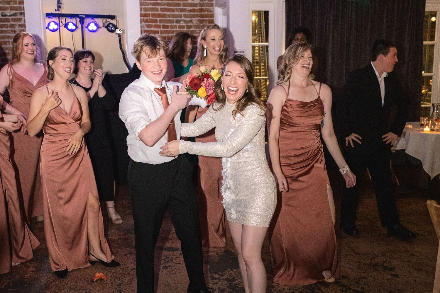 Wedding reception guests dancing and mingling in a brick-walled venue with string lights.