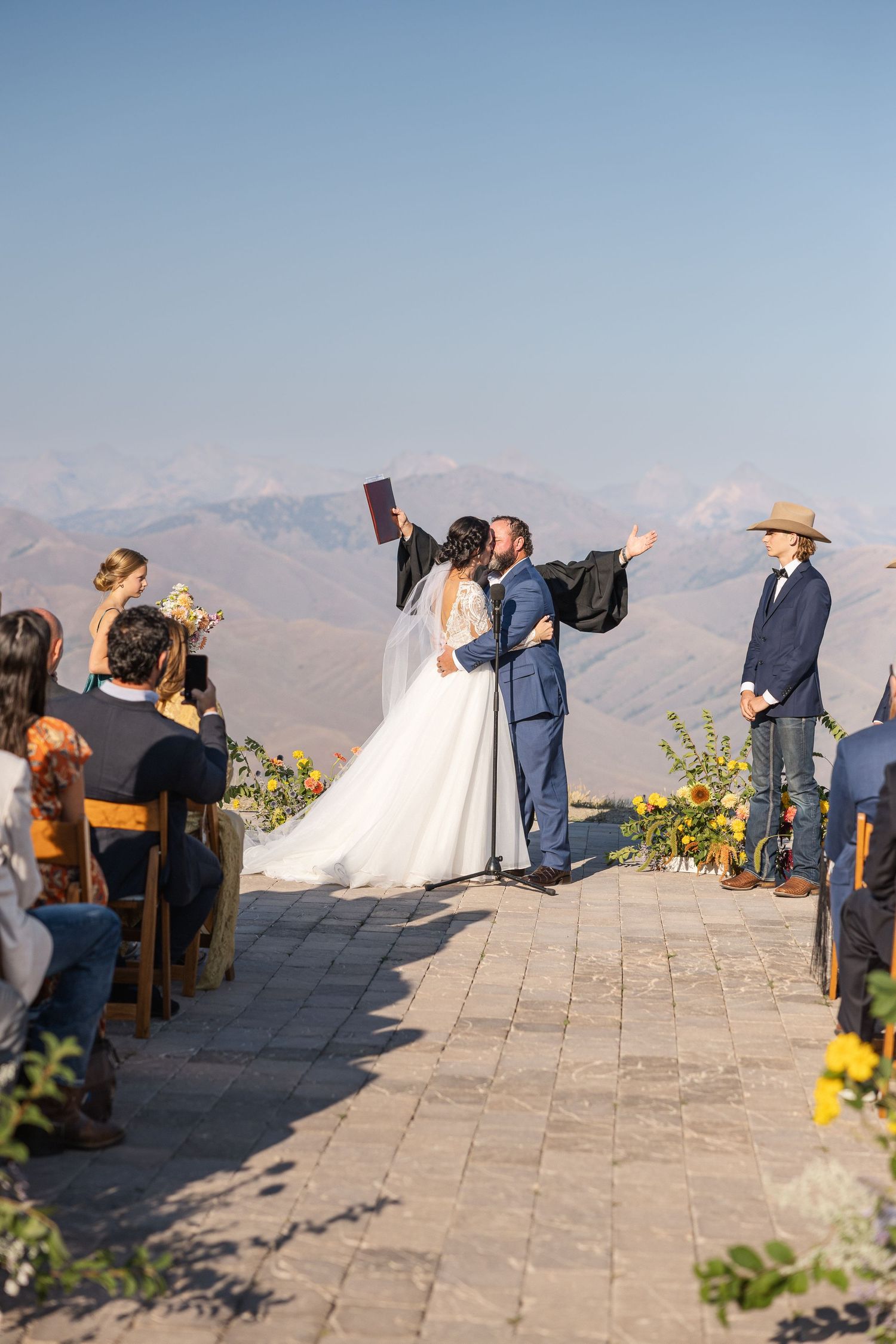 Wedding ceremony guests seated outdoors on a stone patio with mountain views in the background.