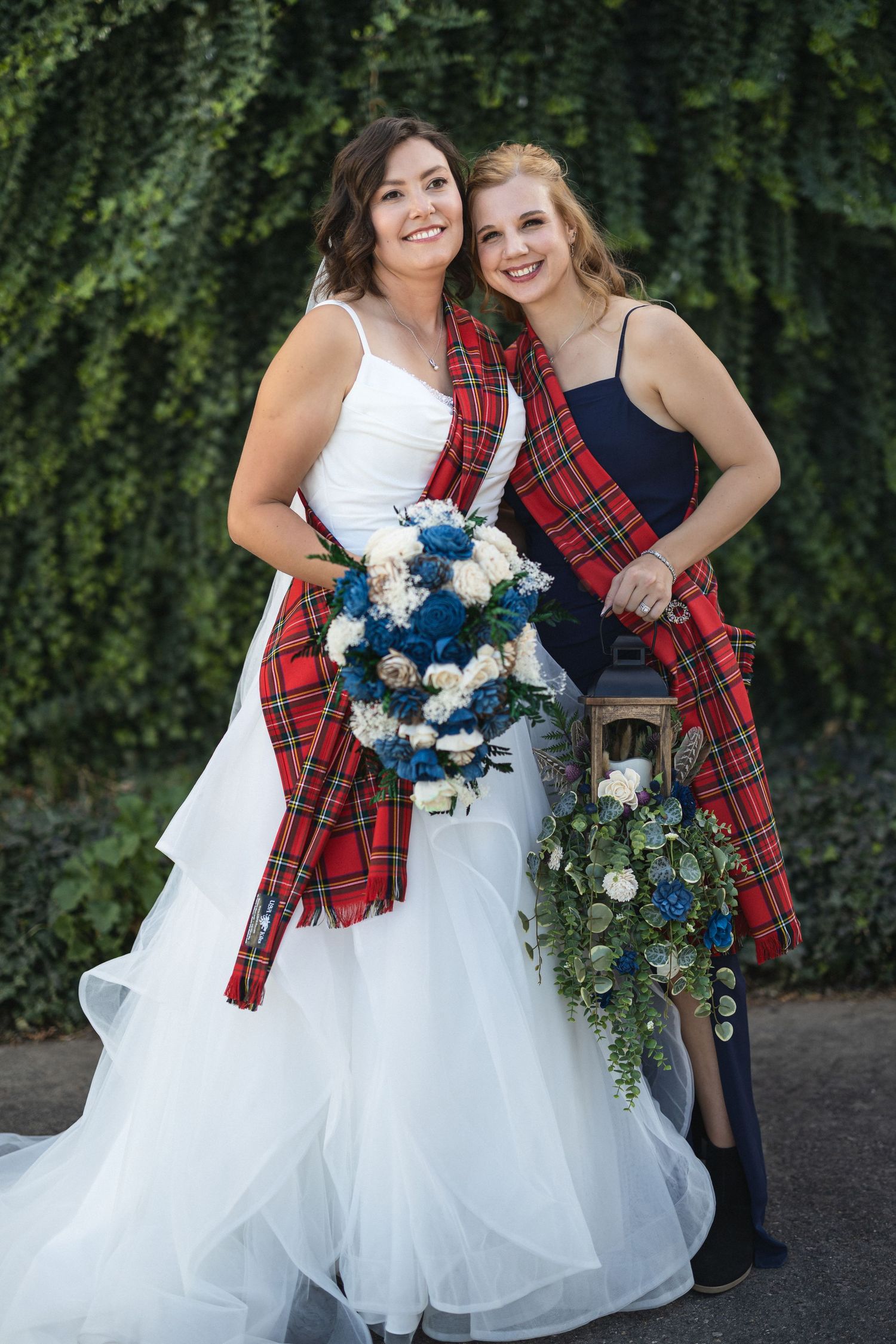 Bride in white gown and tartan sash poses with bridesmaid against ivy-covered wall.
