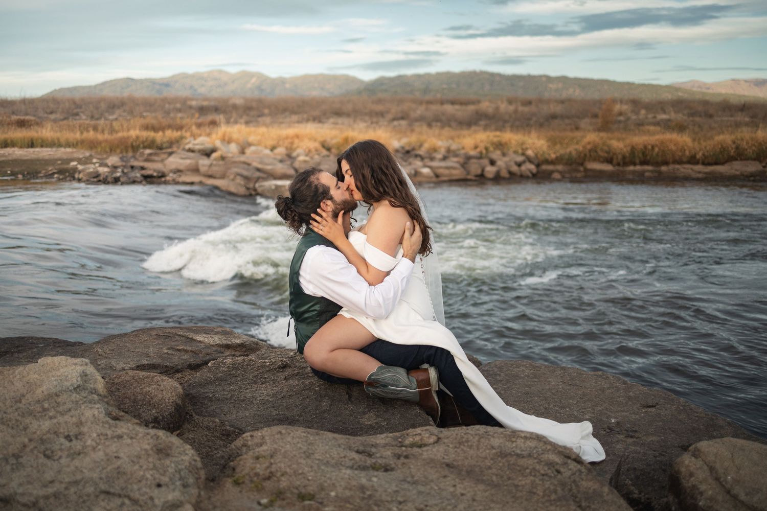 A romantic couple embraces on rocks by a flowing river during their wedding photoshoot.