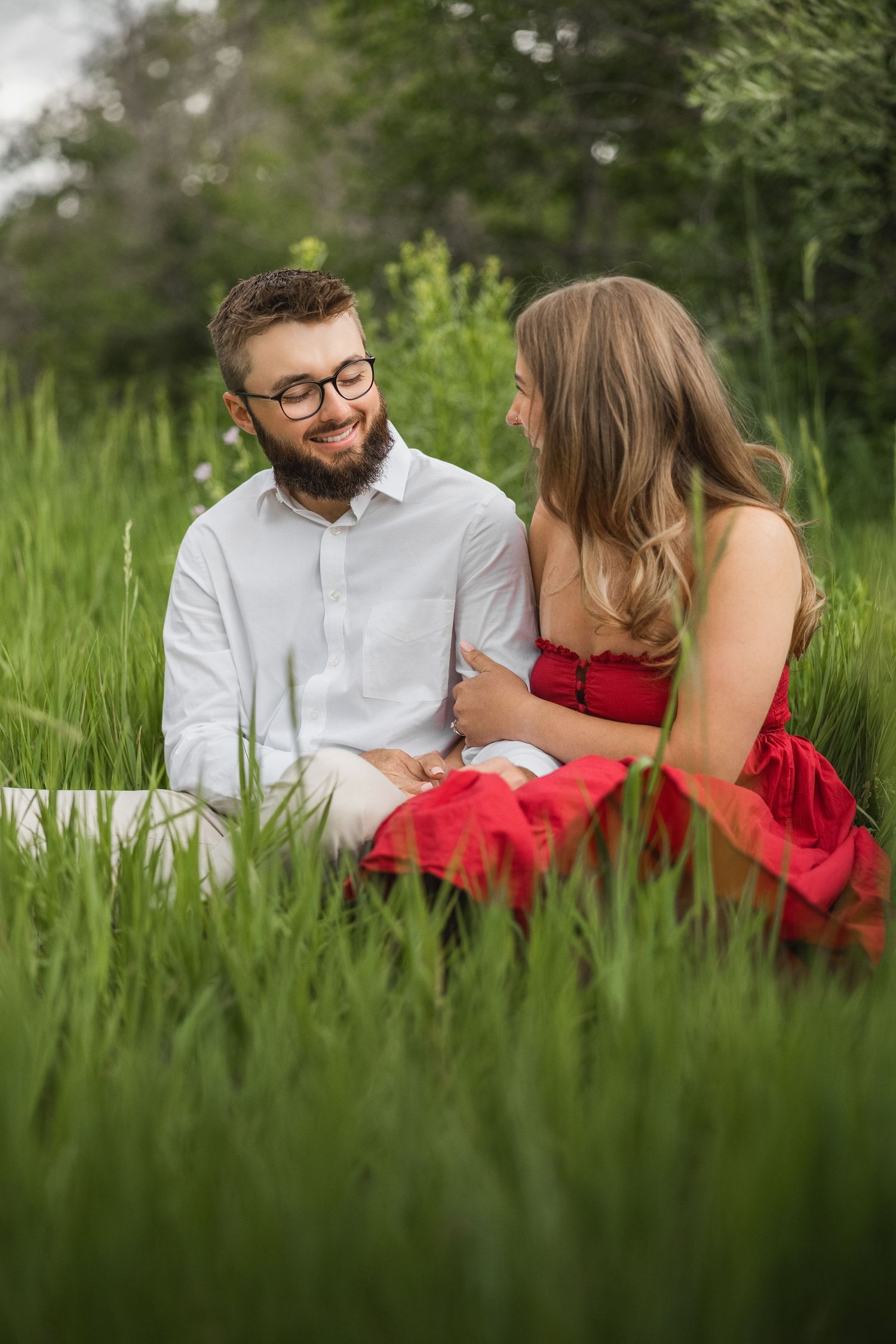 A couple sits intimately together in tall summer grass, with her in a red dress and him in a white button-down shirt.
