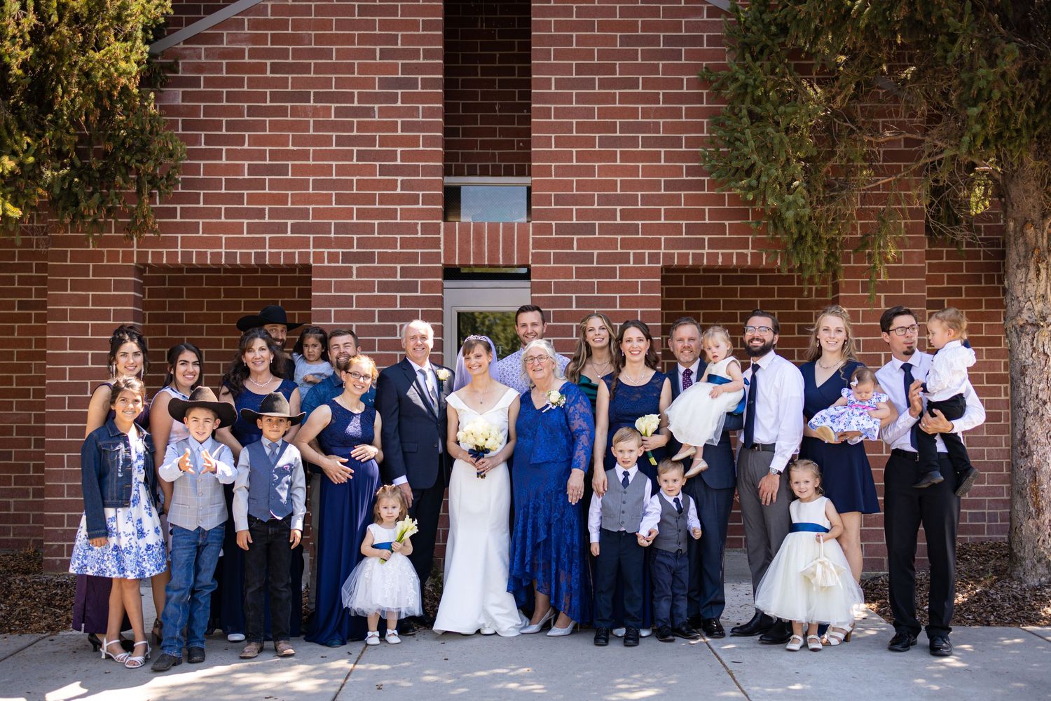 Large wedding group photo with family and children in formal attire against brick backdrop.