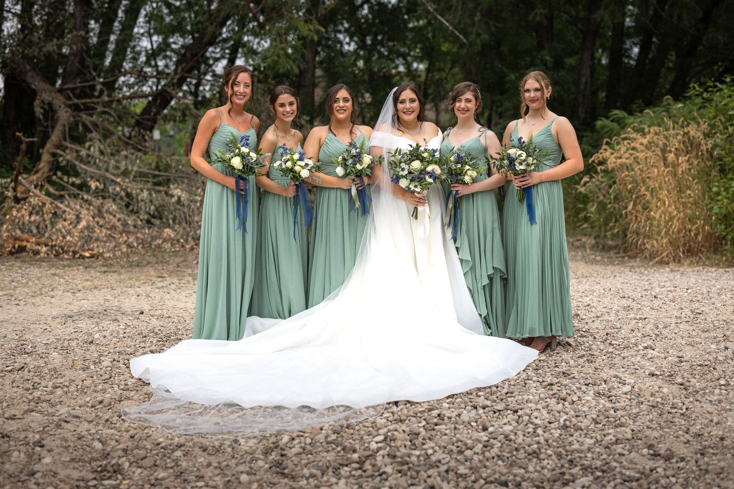 Bride in white gown poses with bridesmaids wearing sage green dresses holding floral bouquets.