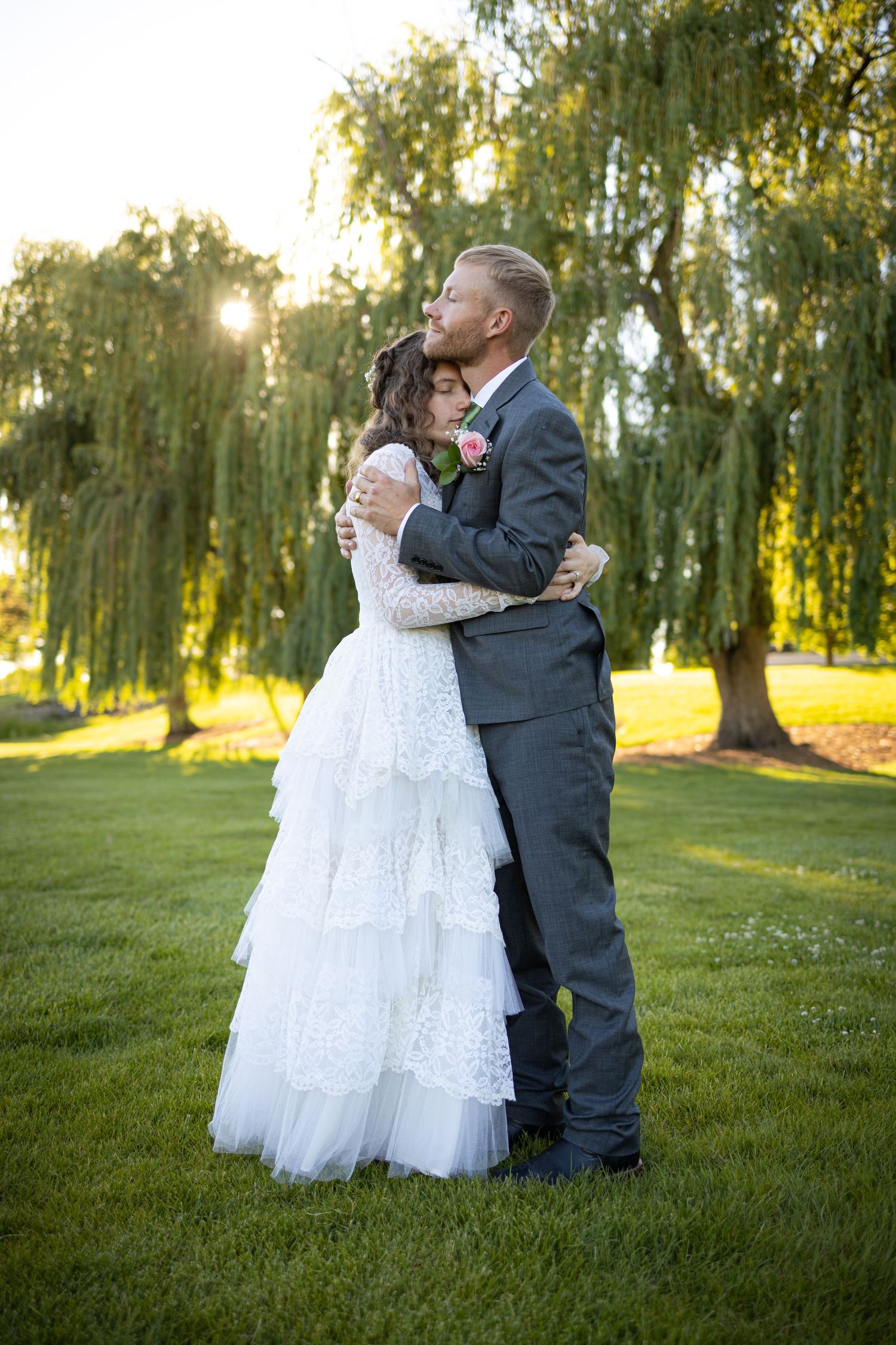 Loving embrace between wedding couple with sunlit willow trees in background.