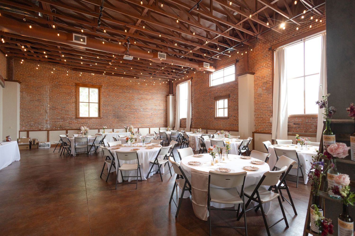Wedding reception hall with string lights, exposed brick walls, and round tables set for dining.