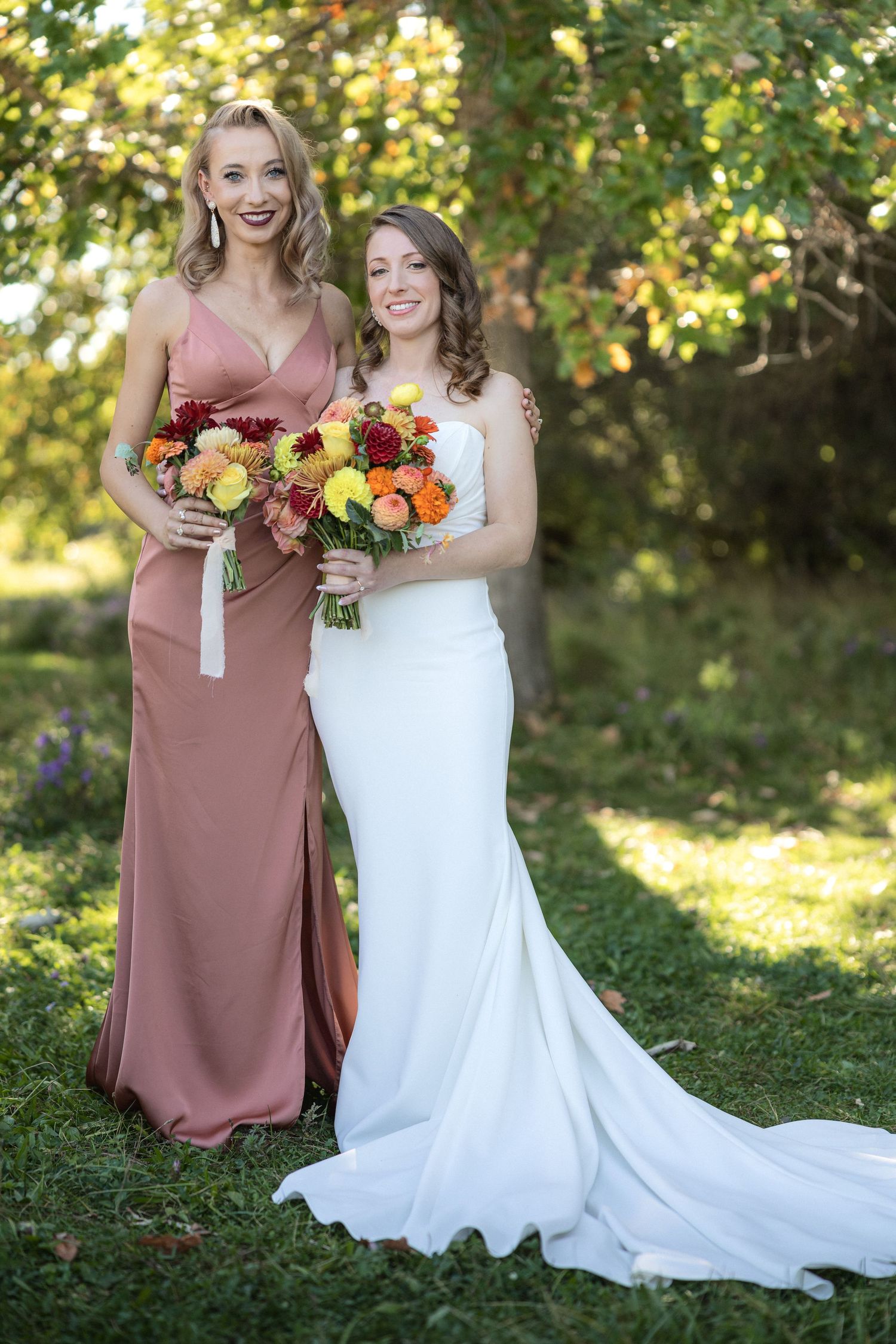 Bridesmaids in rose-colored dresses hold autumn-toned bouquets next to bride in white gown outdoors.