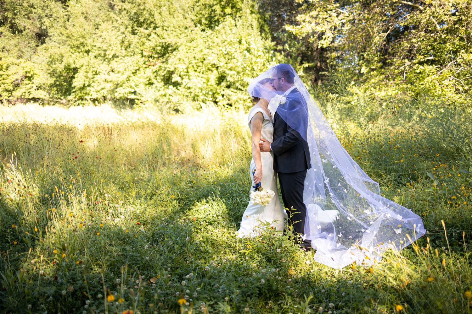 Wedding couple embraces in sunlit meadow with long flowing veil trailing behind.