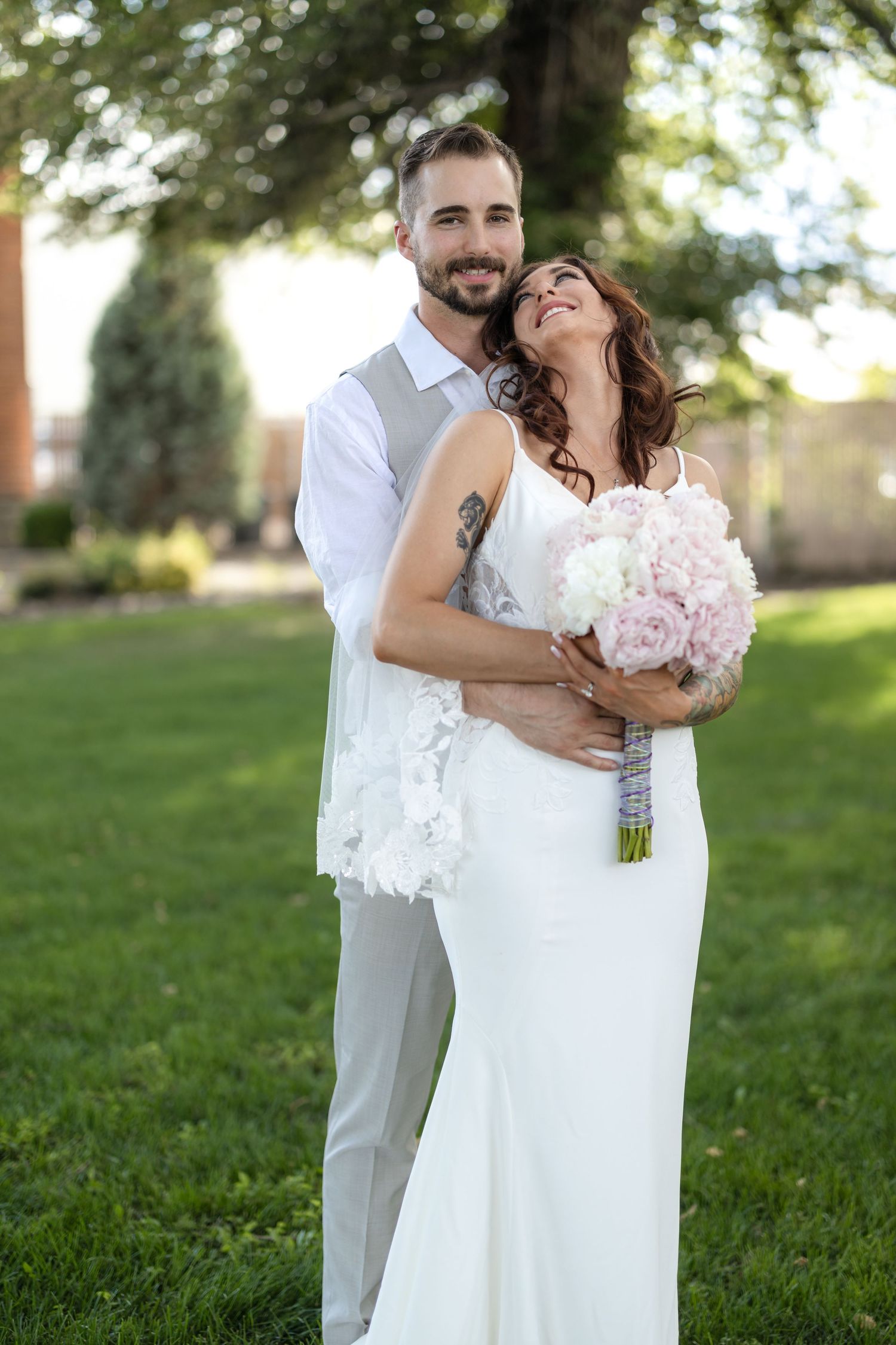 Newlyweds embrace and laugh together on grassy lawn during wedding photoshoot.