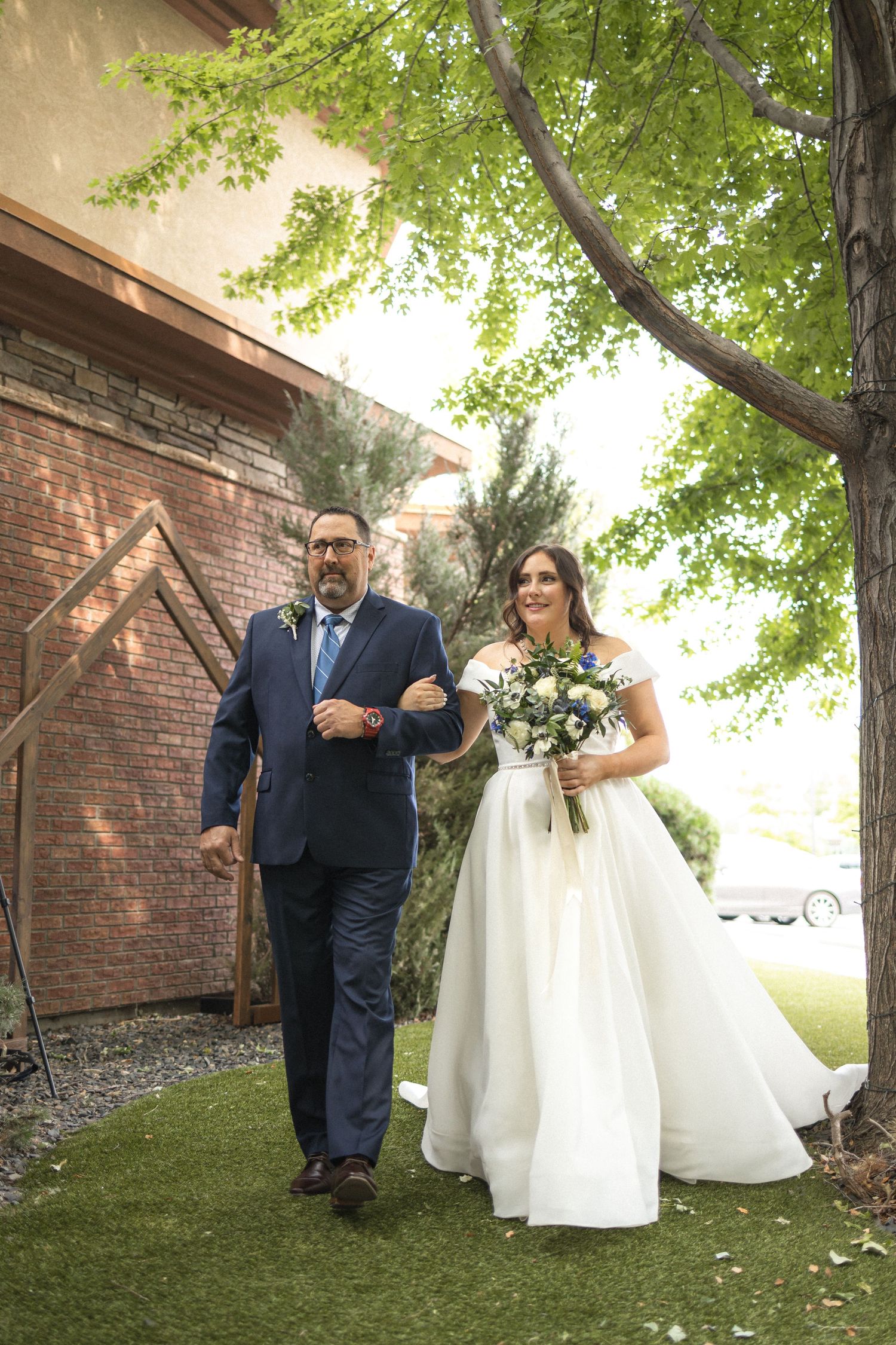 A couple walks arm in arm during a wedding processional against a brick building with overhanging trees.